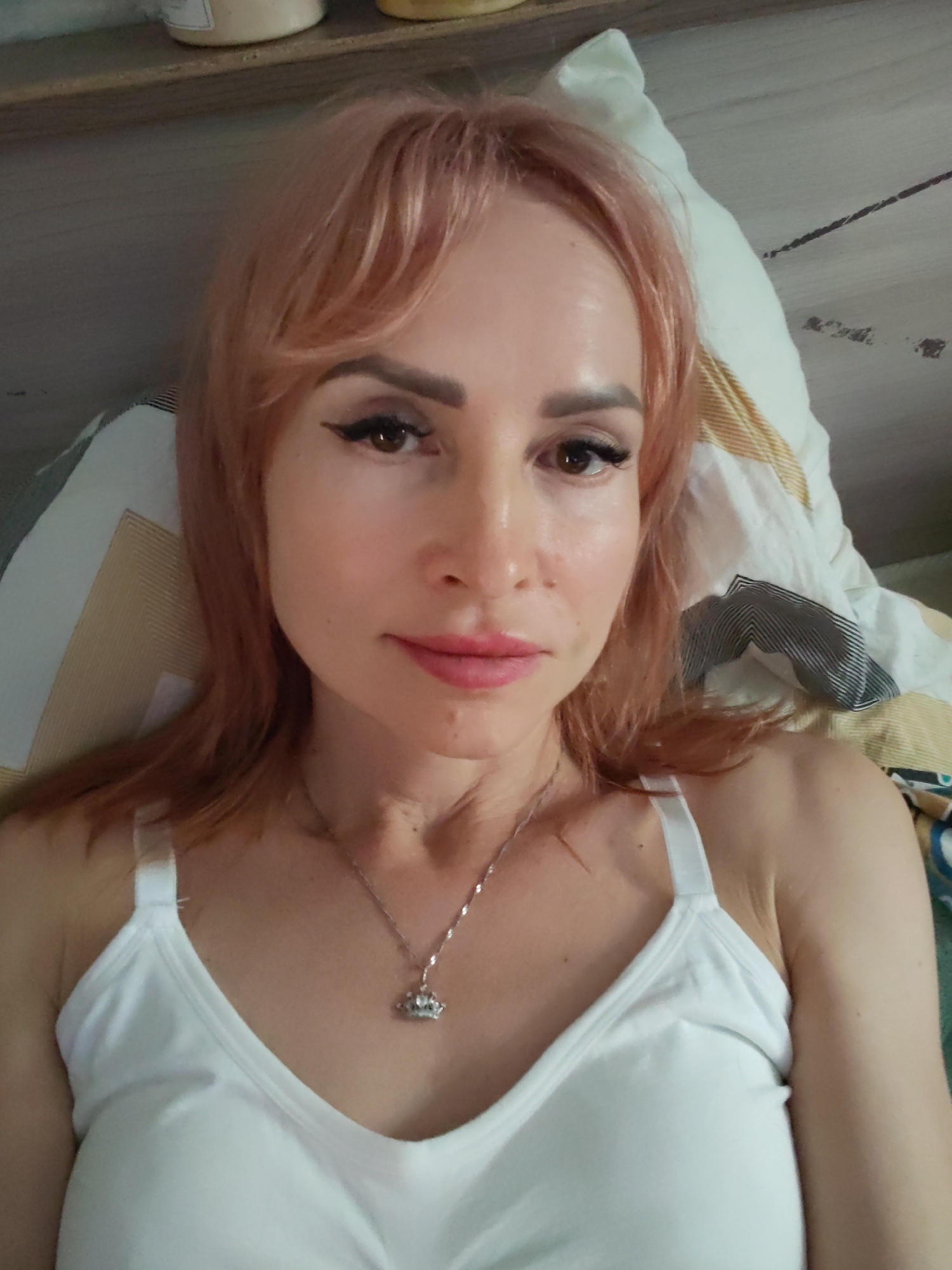 A medium shot selfie of a woman with light pink hair and light skin is taken from a slightly high angle. She is lying on a bed with a white pillow and striped bedding. Her eyes are brown, and she is wearing winged eyeliner and mascara. Her lips are painted a rosy pink. She has a thin silver necklace with a crown-shaped pendant. She is wearing a white tank top. The background shows a shelf with jars, suggesting a domestic setting. The lighting appears to be natural, indicating it might be daytime.