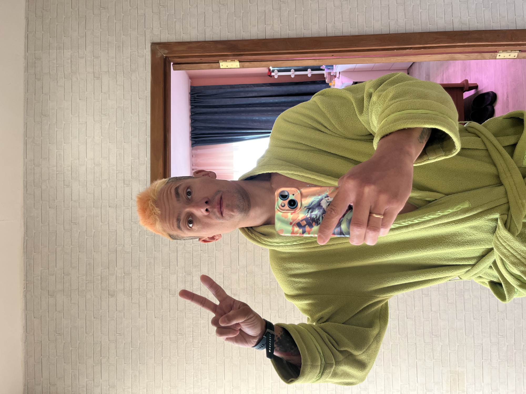 A man with orange-blonde hair is taking a selfie in a mirror. He is wearing a green robe. He is holding a phone with a colorful case, showing a peace sign with his left hand, and wearing a watch. The reflection shows a room with blue curtains, a window, and what appears to be a shelf or table with objects on it. The door frame is wooden. The wall behind the man has a faux brick pattern. The location is presumably Chaikovsky, Russia.