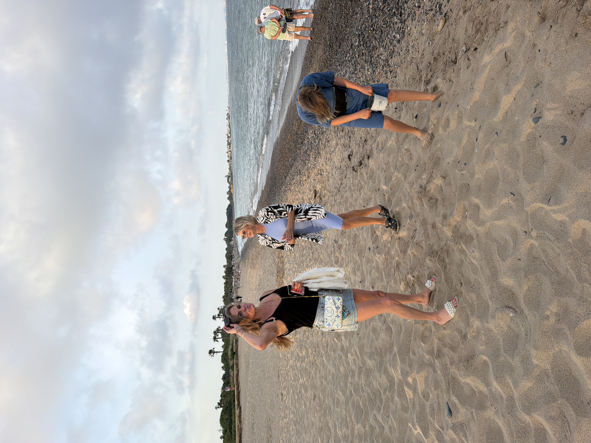 The image shows a beach scene in Mont-roig del Camp, Spain. There are four people visible in the image, three women, and two men. The closest woman, standing on the sand, is wearing a black tank top and denim shorts with a decorative bag. She's wearing sandals and has sunglasses on her head. The second woman, farther away, is wearing a blue outfit with a zebra-striped jacket and black shoes. A third woman is bent over, wearing a blue outfit and holding a cylindrical object. Finally, two men are standing near the water's edge, closer to the waterline. The scene includes sand, the shoreline of the sea, with pebbles and small waves. The sky is overcast with clouds. Trees are visible along the coastline behind the closest women.