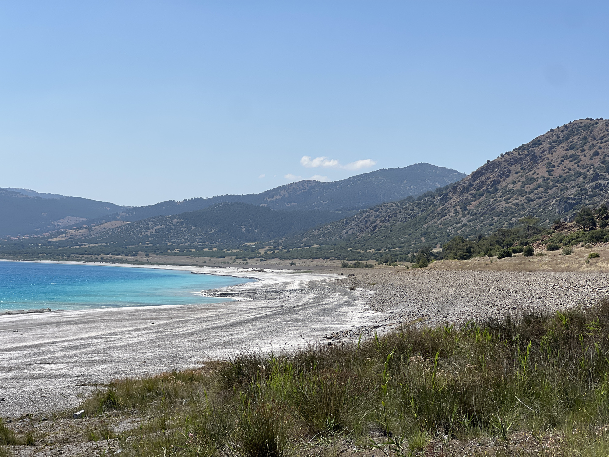 The image shows a wide shot of Salda Lake, a crater lake located in Yeşilova, Turkey, on a clear, sunny day. The lake's water is a vibrant turquoise color, contrasting with the dry, rocky shoreline. The foreground consists of dry grass and scrub brush. Beyond the beach, the landscape rises into rolling, arid hills covered in sparse vegetation. The sky is a bright, cloudless blue.