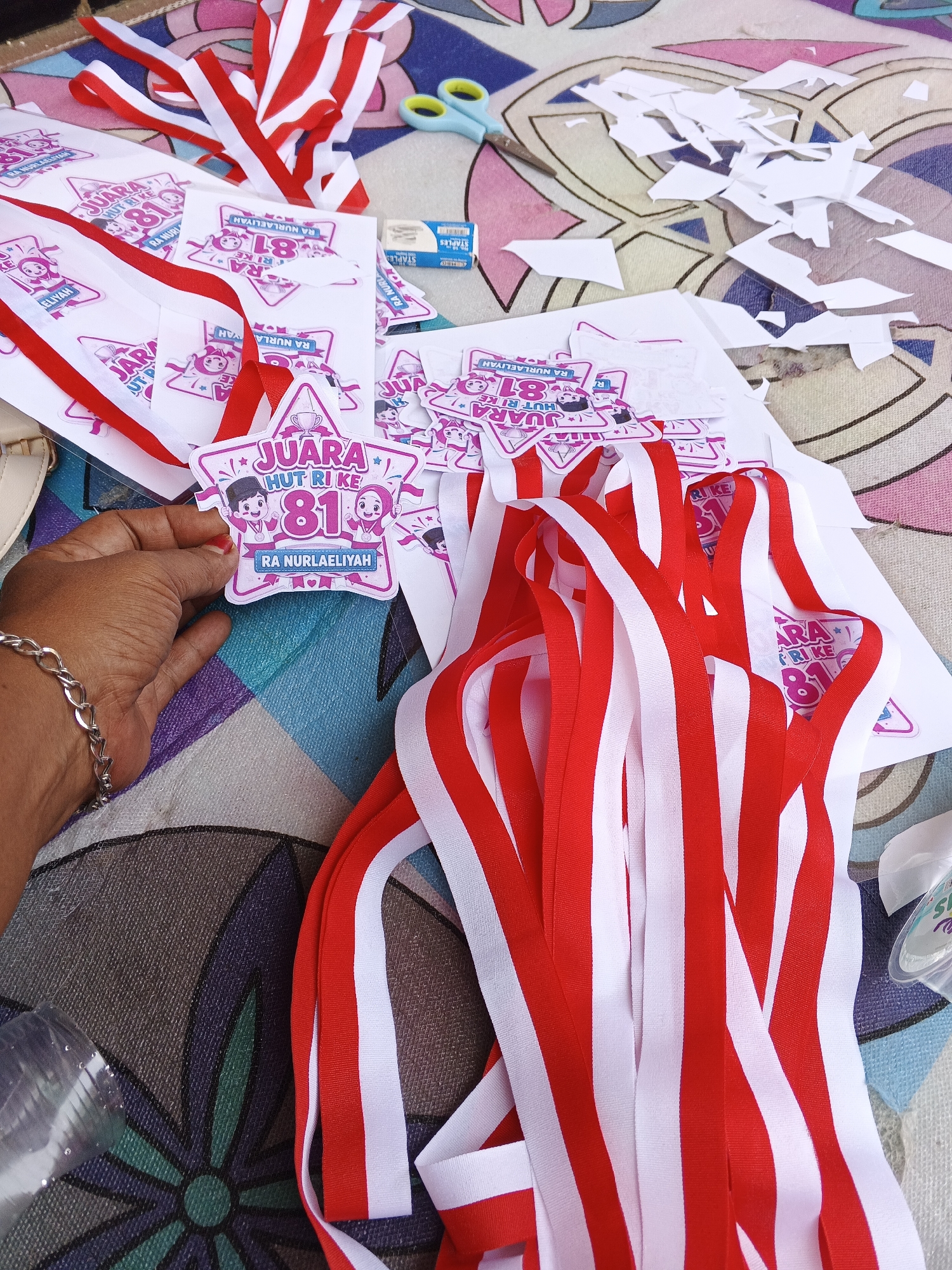 The image shows a close-up of a table covered with a patterned cloth. On the table are various items related to a celebration or award ceremony. Dominating the foreground are numerous red and white ribbons, some bundled together, others loose. In the center, a hand wearing a silver bracelet is holding a star-shaped award. The award features cartoon characters and text that appears to be in Indonesian, including "JUARA HUT RI KE 81" and "RA NURLAELIYAH". Scattered around are cut-out paper shapes, possibly remnants of the award-making process. To the left, a pair of light blue scissors and a small white eraser are visible. In the background, more awards and red and white ribbons are spread out, along with some white paper scraps. The overall impression is one of a craft or preparation area for a local event, likely related to Indonesia's Independence Day ("HUT RI"). The items suggest this scene is taking place in Warungkiara, Indonesia, possibly a school or community center where these awards are being made.