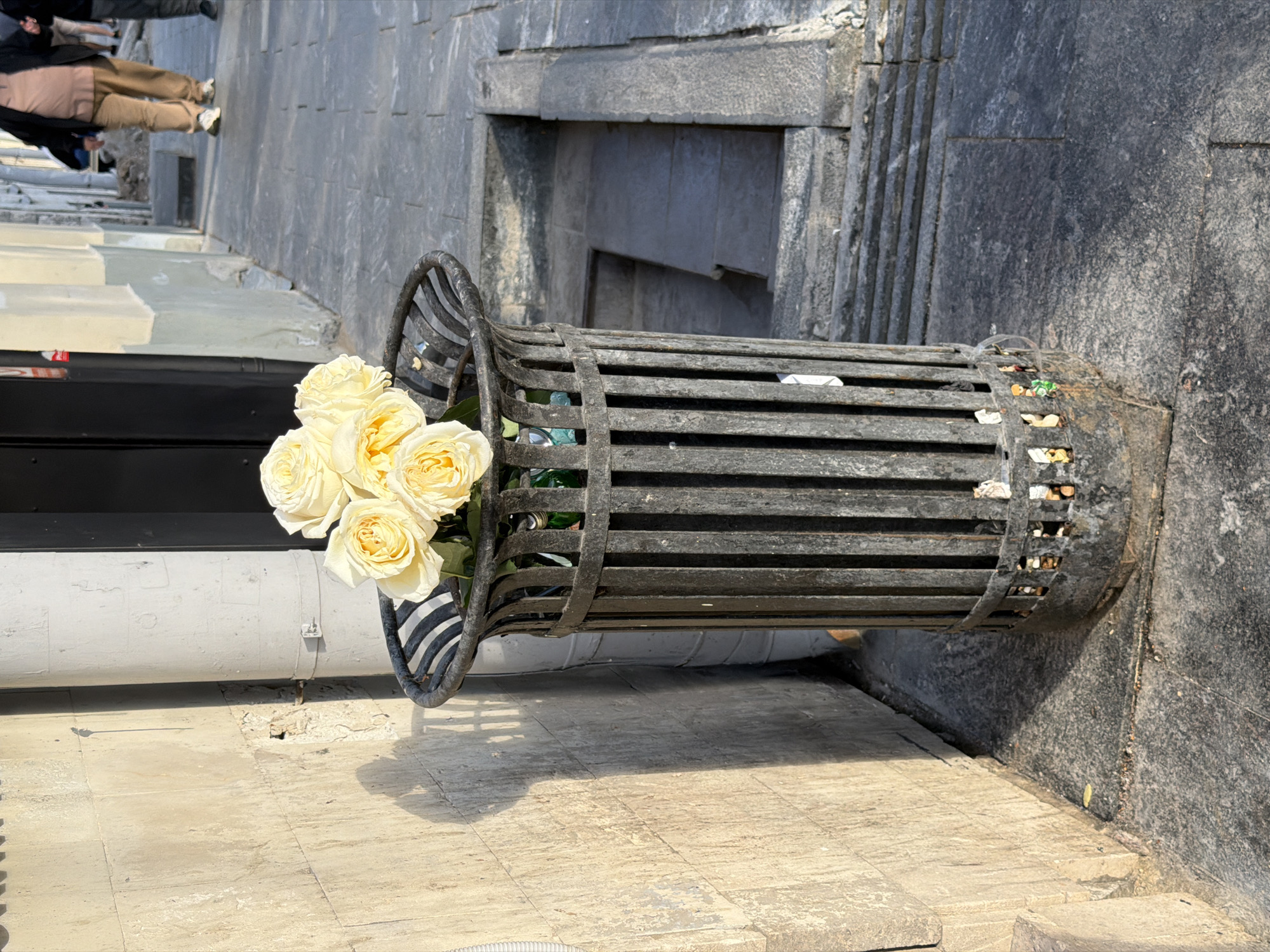 A horizontal, medium shot of a gray, metal, slatted trash receptacle in an urban location in Moscow, Russia, contains a bouquet of light-yellow roses. The trash container is located on a street and a wall is to the right of the receptacle. Inside of the container, below the roses, are items of trash. A white tube and some pavement are to the left of the trash container. In the background are people walking on a gray stone pathway.