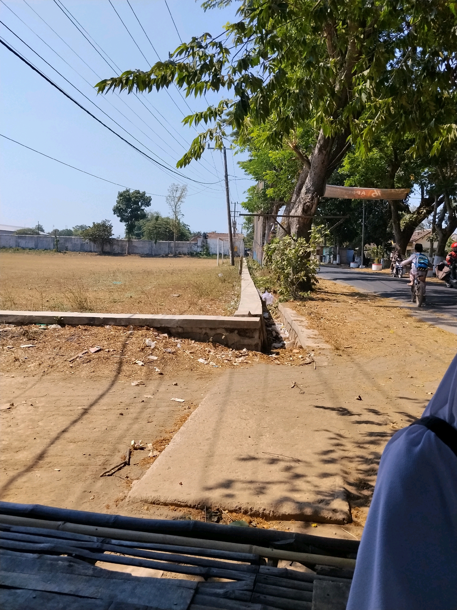 The image depicts a sunny outdoor scene with a street and a field. In the foreground, a person wearing a white headscarf is partially visible on the right side, sitting on a bamboo structure. The ground in the foreground is composed of dirt and a paved walkway, with some debris scattered around.
Beyond the foreground, a dirt field stretches out, bordered by a low concrete wall. A further concrete wall runs along the edge of the field, separating it from the street. Several trees with dense green foliage are present, casting shadows on the ground. One large tree has a banner hanging from it.
The street itself is paved and shows some signs of wear. Several people are visible on the street. One person is riding a bicycle, carrying a blue backpack. Other individuals are on motorcycles in the distance. Electrical wires are suspended above the scene, supported by utility poles. The sky is clear and blue. In the background, there are buildings and more trees, suggesting a semi-urban or rural environment within the city of Jambeanom, Indonesia. The overall impression is of a typical day in this location.