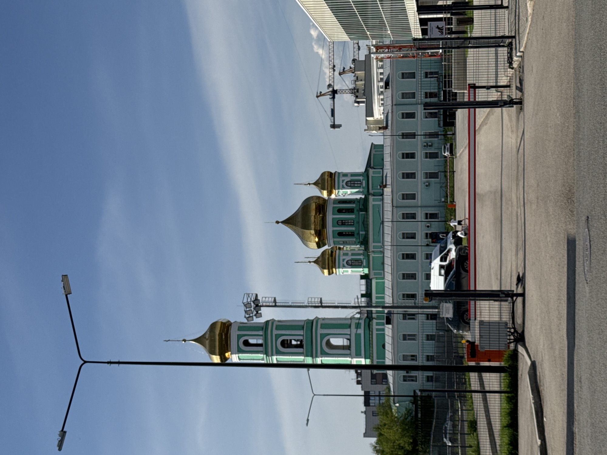 The image shows the Holy Trinity Cathedral in Perm, Russia. The cathedral has three prominent golden domes. It stands adjacent to a modern building, possibly an office or residential complex, and construction cranes are visible nearby. A parking lot with vehicles is next to the building. A street and lampposts are in the foreground. A black iron fence separates the cathedral from the road. The sky is clear and blue.