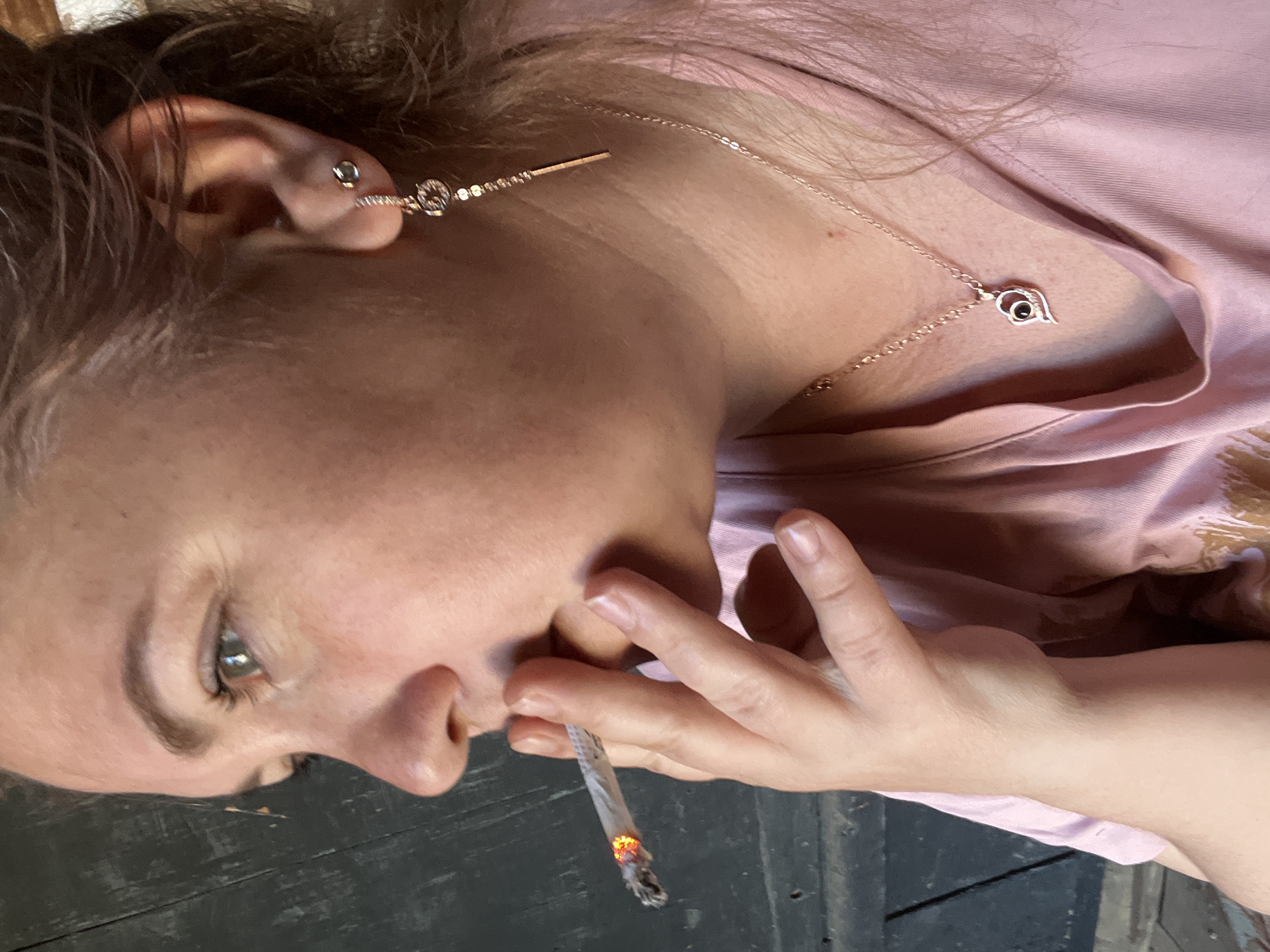 The image shows a close-up, side profile of a woman smoking a cigarette. She has blonde hair and is wearing a pink shirt, a necklace with a heart pendant, and earrings. The background appears to be a dark, weathered wooden surface. There are no discernible elements in the image that would indicate a specific location within the city of Фоки, Russia.