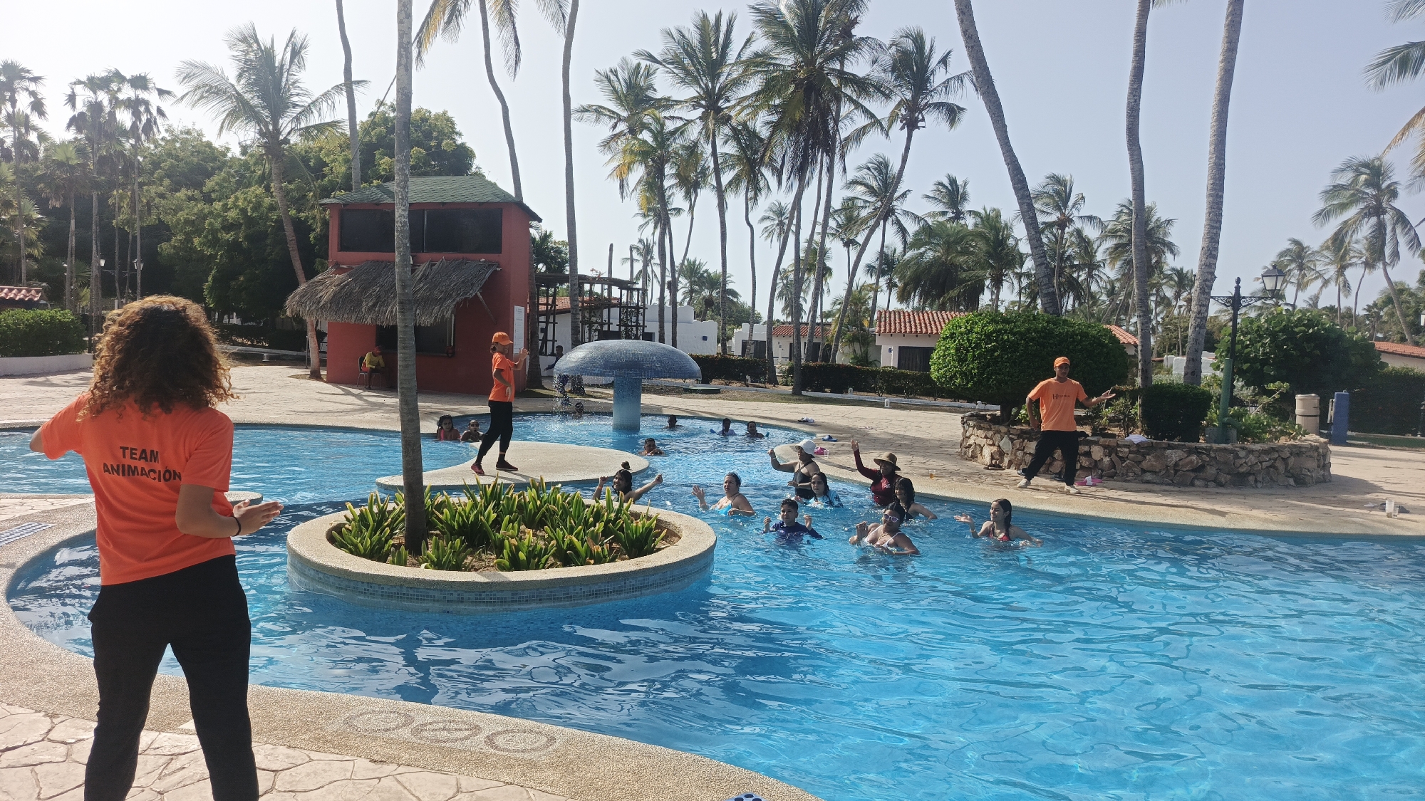 The image captures a lively scene at a swimming pool in Playa El Agua, Venezuela. A group of people are participating in a water aerobics class. Two instructors wearing bright orange shirts labeled "TEAM ANIMACION" are leading the class, one standing on a raised circular planter and the other standing on the side of the pool.
The pool is surrounded by palm trees and various buildings, including a red building with a thatched roof. A mushroom-shaped structure is also visible in the pool area. The pool is filled with clear blue water.