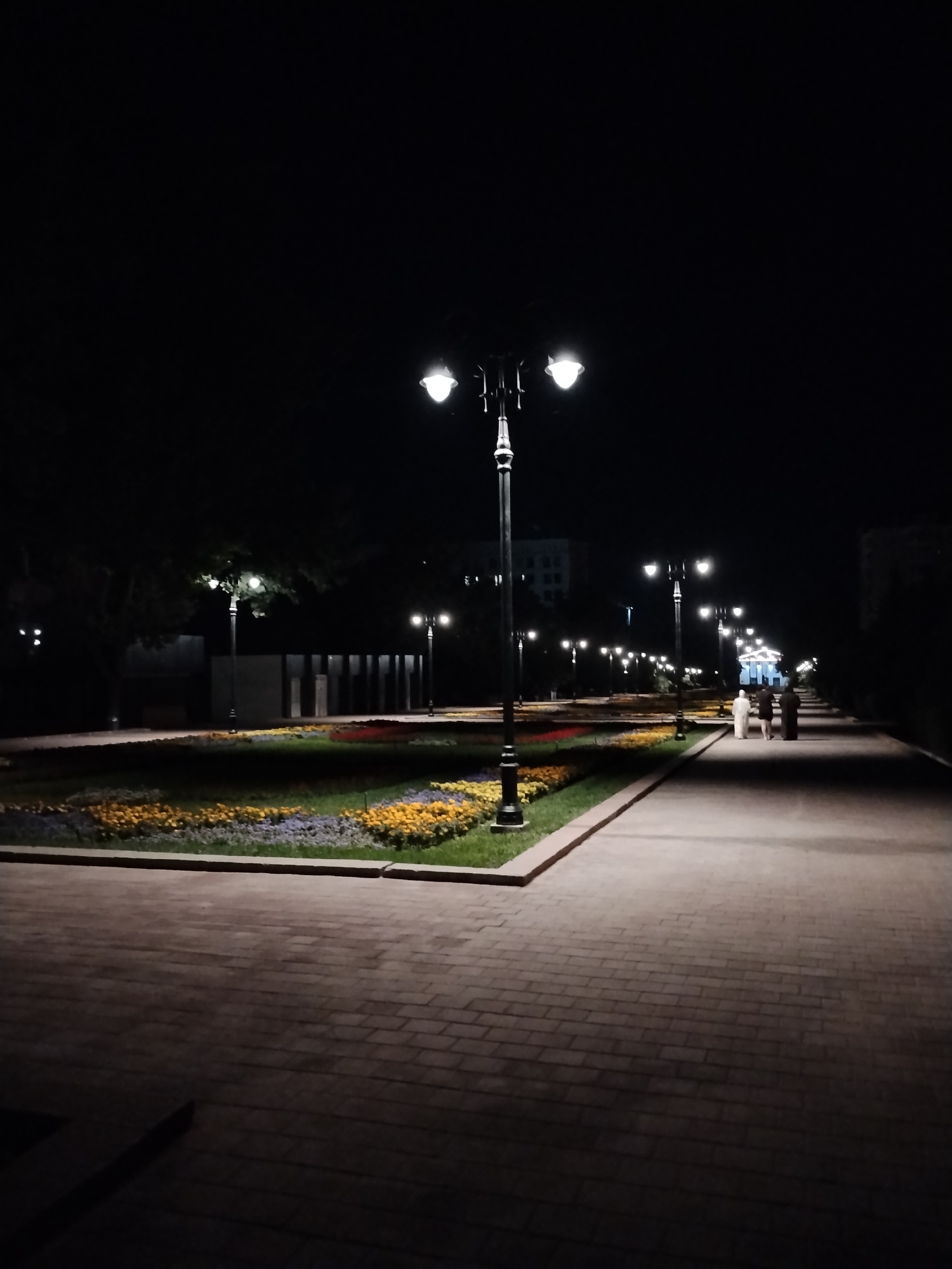 The image depicts a paved walkway in a park at night, illuminated by streetlights. The walkway is bordered by flowerbeds containing yellow and purple flowers. Several lampposts are visible along the walkway, casting light on the path and the surrounding area. In the background, there are trees and a building. Three individuals, two in light-colored clothing and one in darker attire, are walking away from the camera on the walkway. The scene is consistent with a public park in Bishkek City, Kyrgyzstan, likely an area designed for evening strolls.