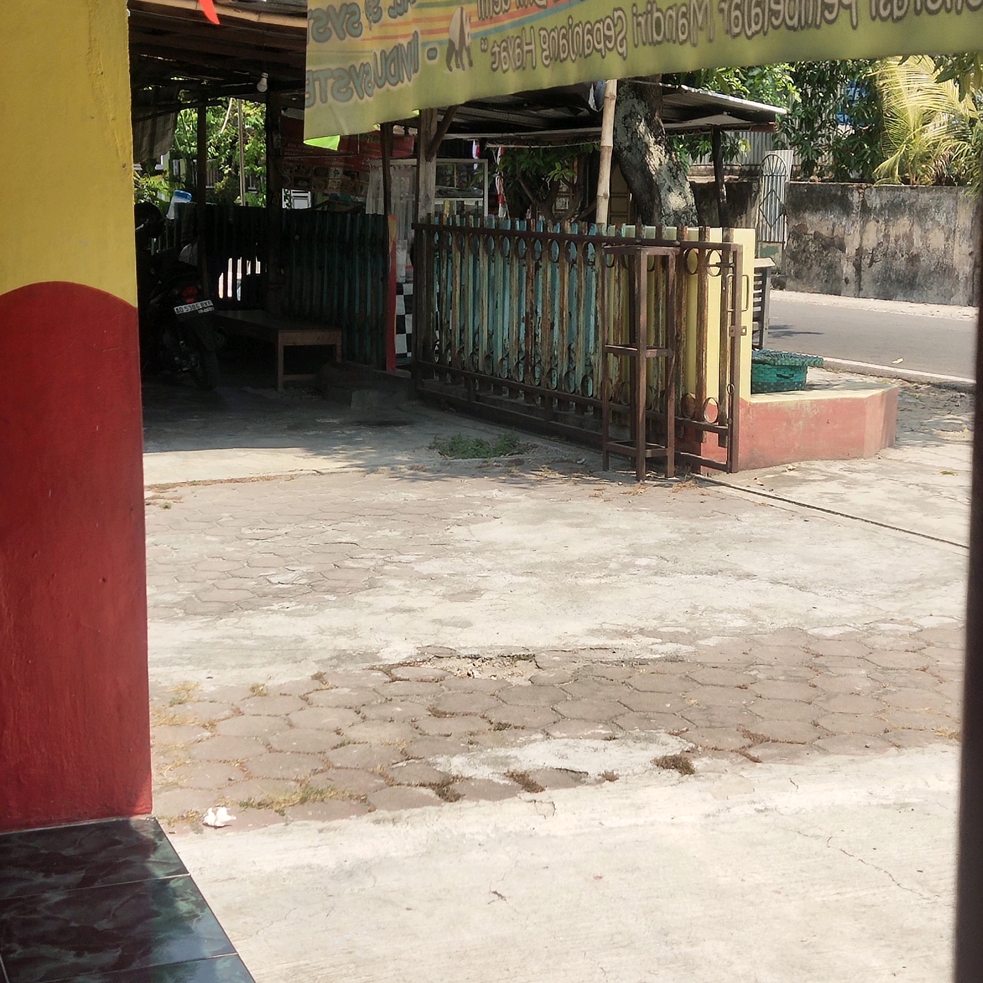 The image captures an outdoor scene in Sragen, Indonesia, likely a street-side area in front of a business. A yellow and red painted wall forms the left edge of the frame, with dark, glossy tiles on the ground in the lower left. In the middle ground, a weathered, multi-colored metal fence with vertical bars and decorative scrollwork is visible, opening to a paved courtyard. Beyond the fence, a motorcycle with a license plate reading "AD 5305 BXC" is parked, and a simple wooden bench sits in the shade. A large tree trunk with textured bark is prominent behind the fence. A section of a building with a corrugated metal roof and a sign with blurred text is also present. The ground is paved with interlocking hexagonal concrete blocks, some of which are uneven or broken, with patches of dirt and dry grass. In the background, a concrete wall runs along the edge of a paved road, suggesting a commercial or residential area. The overall impression is one of a moderately used, somewhat aged urban environment.