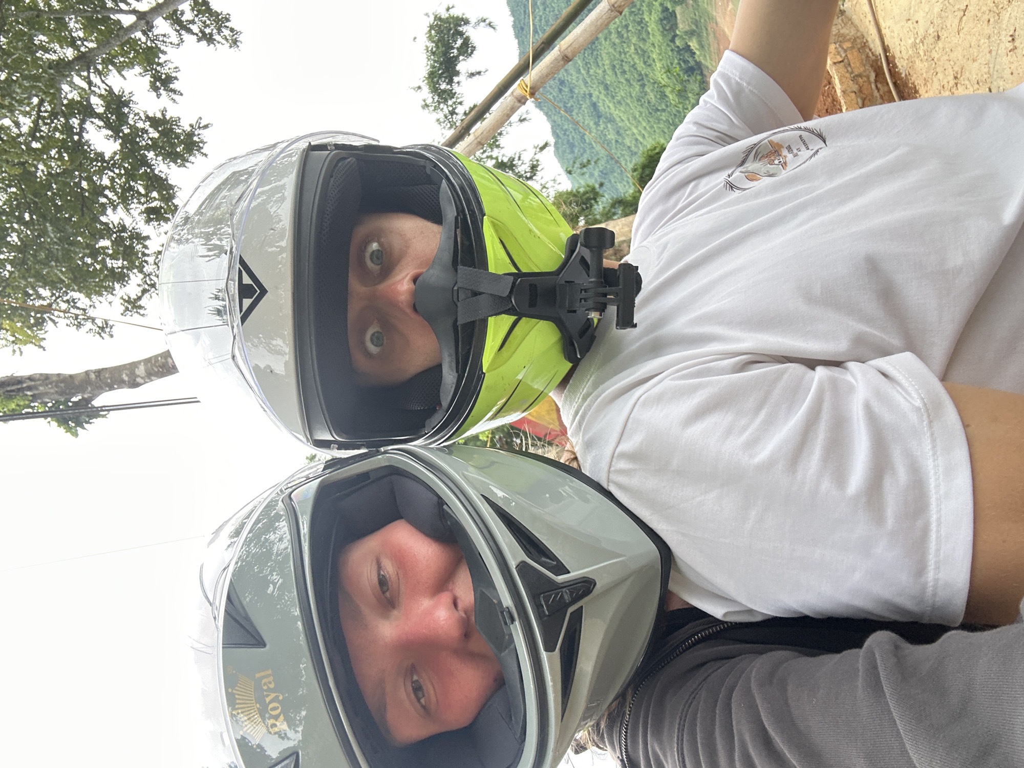 Two individuals are positioned in the foreground, both wearing full-face helmets, in what appears to be an outdoor setting. The person on the left is wearing a light grey helmet with a gold crown emblem and the word "Royal" visible. Their face is visible through the helmet opening, showing fair skin flushed with red, brown eyes, and reddish-brown hair. They are wearing a dark grey jacket.
The person on the right wears a bright lime green helmet. Their face is also visible, with wide, surprised-looking eyes and a pale complexion. A black chin guard with a GoPro mount is attached to their helmet. They are wearing a white t-shirt with a graphic design on the left chest area.
The background suggests a rural or mountainous environment. Lush green hills, dense vegetation, and trees are visible. Several bamboo poles are propped up in the middle ground, one of which has a yellow string tied to it. The sky is overcast and bright white. The setting is identified as Bản Phúc, Vietnam. The overall impression is a selfie taken by two people on a motorcycle, possibly during a scenic journey.