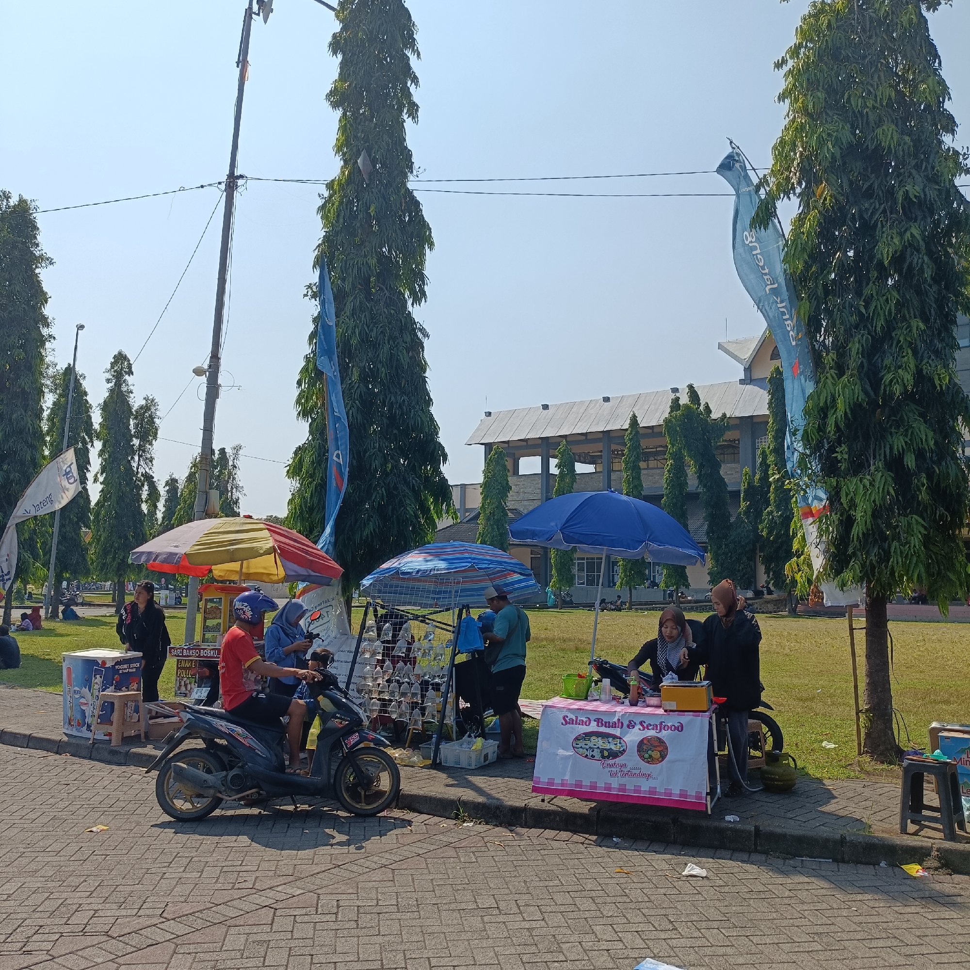 The image depicts a street scene in Kendal, Indonesia, characterized by an outdoor market or food stall area. In the foreground, a paved walkway is lined with parked motorcycles and several food vendors. One vendor, a woman wearing a hijab, stands behind a stall selling "Salad Buah & Seafood" which is adorned with a pink and white checkered tablecloth and features images of various fruits and seafood. Next to her, another vendor in a black jacket is serving customers. A motorcycle with two people on it is parked near the stalls. In the background, tall trees line a grassy area, and a large building with a modern architectural style is visible. Several flags, some bearing text, are displayed throughout the scene. The overall atmosphere suggests a busy, sunny day at a local gathering or event.