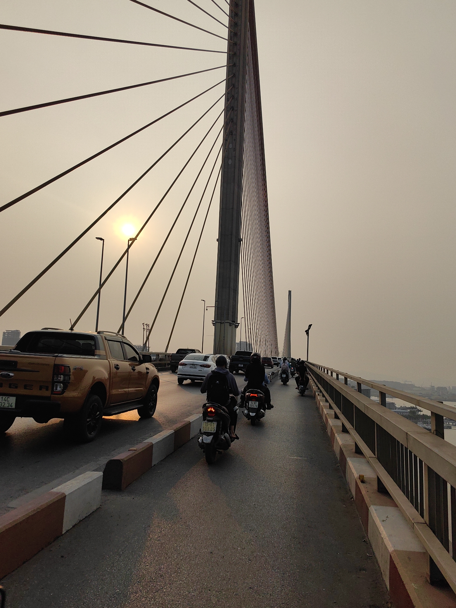 Captured on a bridge in Hong Gai Ward, Vietnam, the image presents a dense scene of vehicular traffic under an overcast sky. A golden Ford Ranger pickup truck dominates the left foreground, with a license plate reading "14C-032.34". The truck is followed by several motorcycles, one displaying a license plate "15-G1 085.03", adding to the congestion. A white sedan and black SUV are also visible further down the road. Overhead, the bridge's suspension cables radiate from a tall tower, with the sun shining through, creating a hazy, golden light. The bridge's railing, composed of vertical bars and a colored concrete base, runs along the right side of the frame, offering a glimpse of the waterway and city skyline in the distance. Overall, the image conveys a sense of bustling activity amidst the urban infrastructure of Hong Gai Ward.
