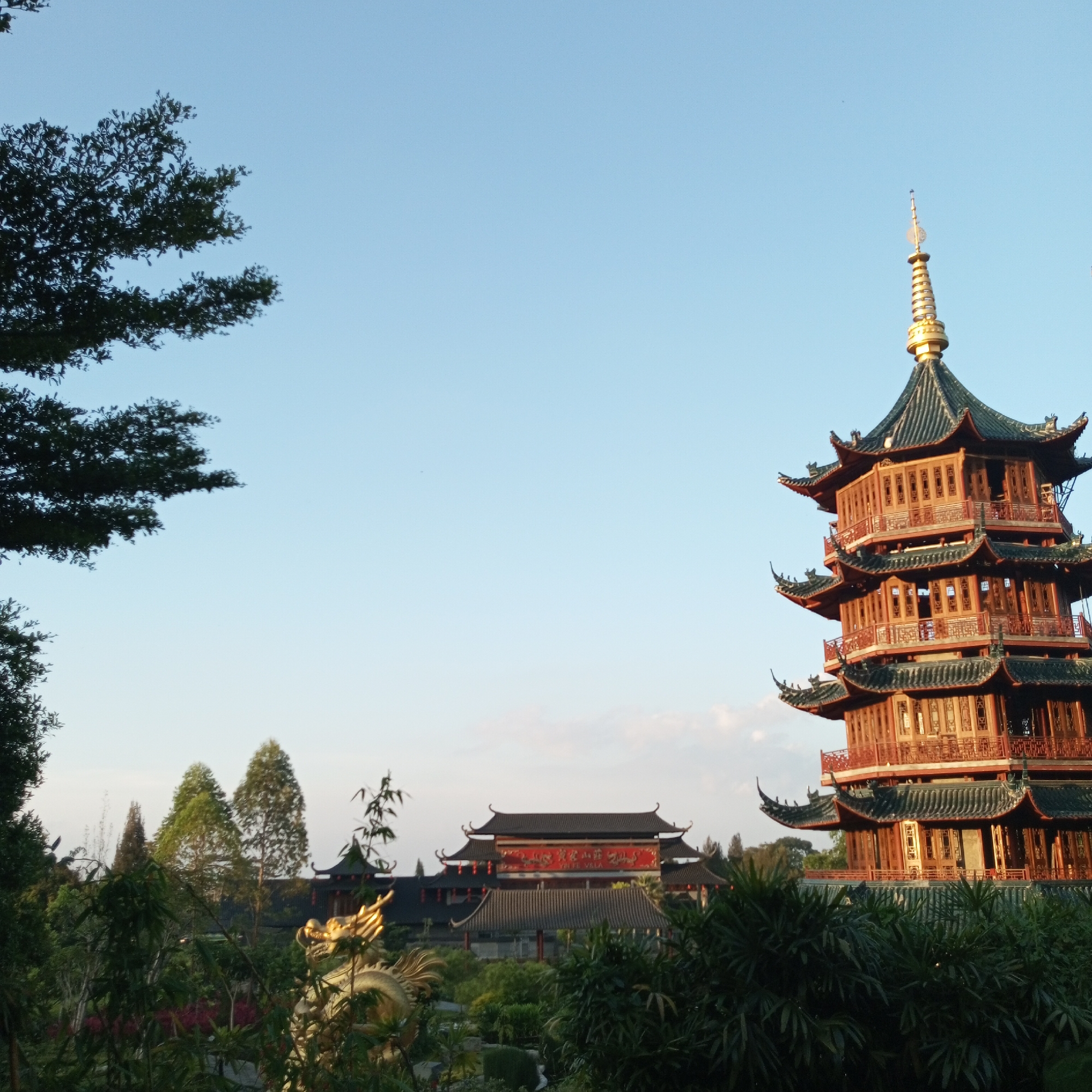 The image shows a multi-tiered pagoda, characteristic of East Asian architecture, situated in the city of Suka Makmur, Indonesia. The pagoda is constructed with dark tiled roofs and vibrant red wooden accents. It is surrounded by lush green foliage and trees, with a clear, pale blue sky as the backdrop. In the middle ground, a traditional East Asian building with a red banner displaying calligraphy can be seen, also featuring dark tiled roofs and red accents. To the left of the pagoda, a golden dragon statue is partially visible, suggesting a decorative element within the landscape. The scene appears to be a park or cultural site, bathed in the soft light of either sunrise or sunset, indicated by the warm tones on the pagoda and the gentle illumination of the sky. No people are present in the image.