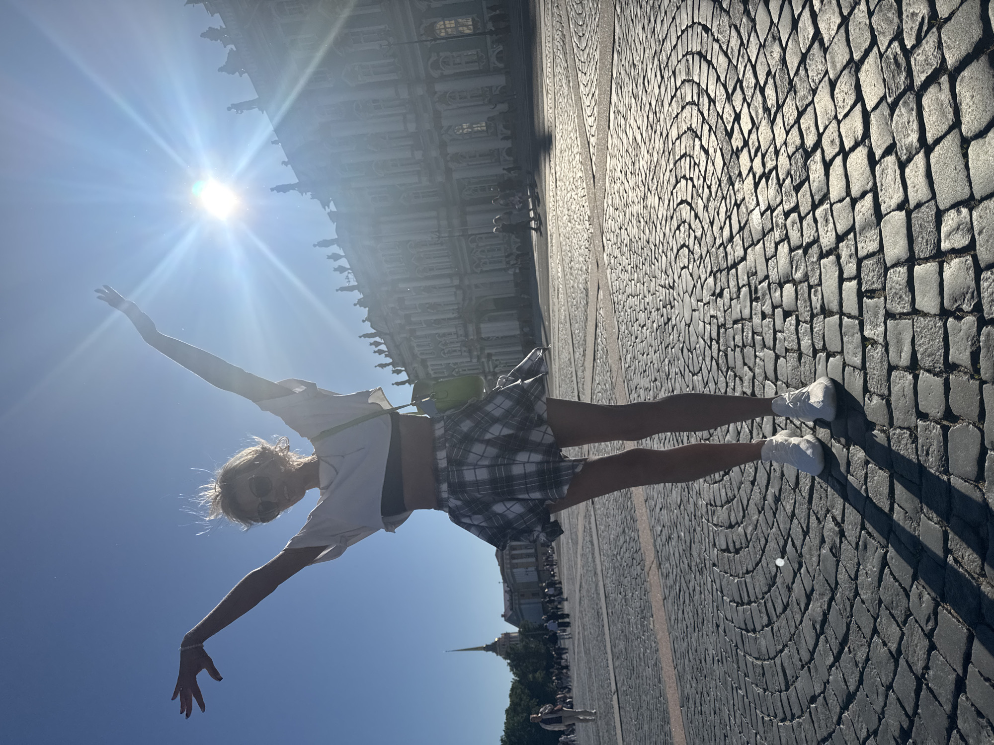 A young woman with blonde hair, wearing sunglasses, a white crop top, a plaid mini-skirt, and white sneakers, stands with her arms outstretched in a cobblestone plaza in Saint Petersburg, Russia. She is facing the camera, with the sun shining brightly behind her. In the background, the grand facade of the Winter Palace is visible, with its many windows and statues. The sky is clear and blue. People can be seen walking in the distance, and a spire of a building is visible to the left. The cobblestones are arranged in a circular pattern. The overall mood of the image is one of joy and freedom.