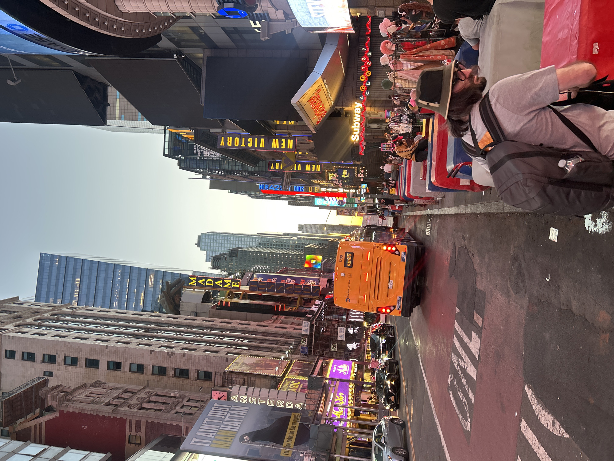 This media file captures a vibrant street scene in New York City, likely Times Square, bustling with activity during what appears to be late afternoon or early evening. The sky is clear and bright, contrasting with the numerous illuminated billboards and neon signs.
In the foreground, a man with a beard, wearing a hat, grey t-shirt, and a dark backpack, is seated on a colorful street barrier, facing left. The street is filled with traffic, including an orange New York City bus with the route "SIM25" and identification "1730" visible on its rear, moving away from the viewer. Several cars follow behind it. The road features a prominent red-painted lane with the partial text "ONLY" (likely for "BUS ONLY").
The buildings lining the street are adorned with an array of large, bright billboards and signs. Visible text includes "NEW VICTORY," "Subway" (the restaurant), "NEW 42ND STREET," "MADAME," "Aladdin," "AMSTERDAM," "Potter" (likely referring to Harry Potter), and "IT AIN'T JUST MANHATTAN" with the tagline "I'm New Yorker, too. Give me respect." Other partially visible texts include "DIOS," "Milly," and various subway line indicators such as "Q," "R," "W," "2," "S," and "C." The scene depicts a lively urban environment, characterized by constant movement, advertising, and a diverse street-level experience.