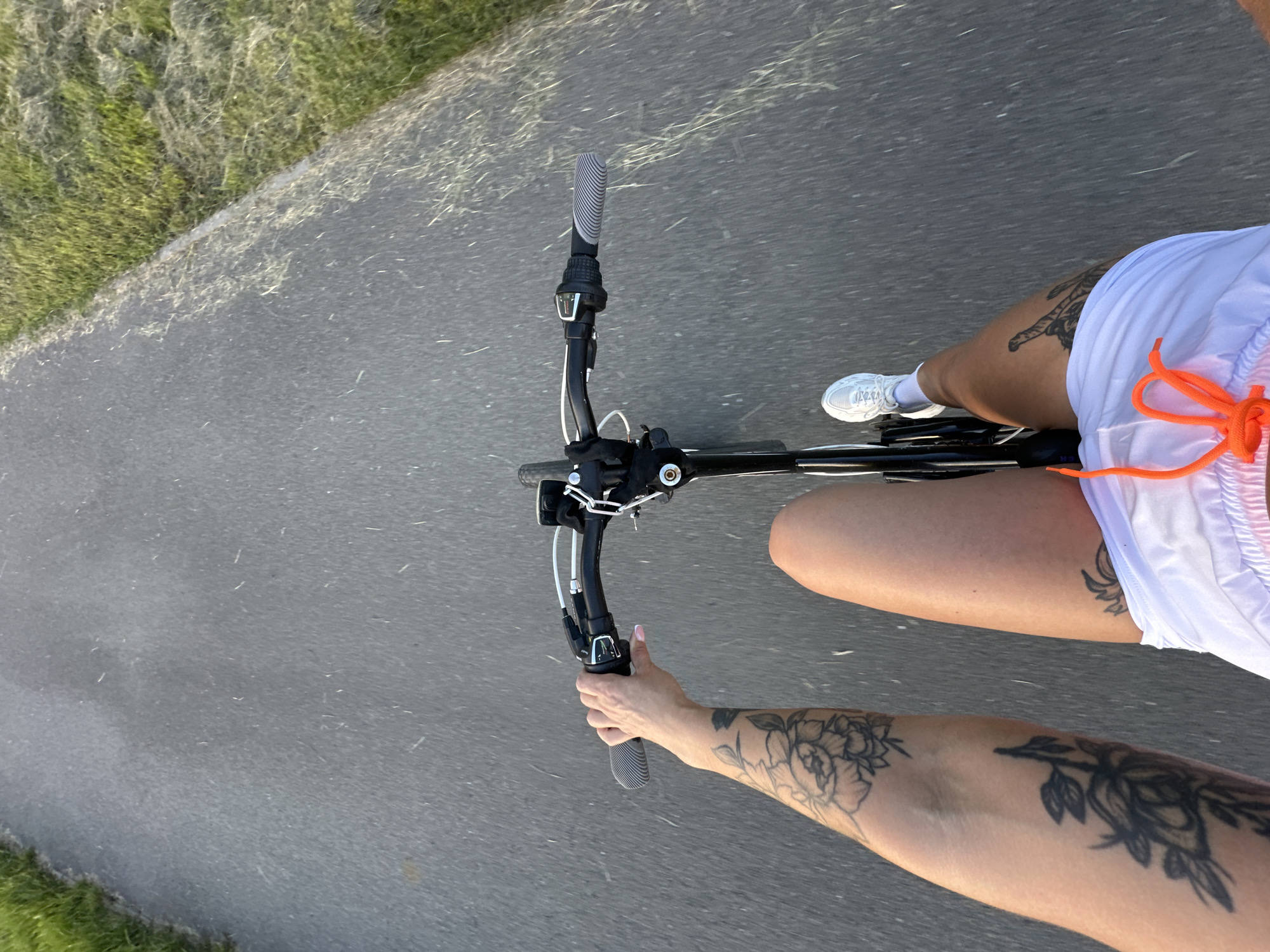 A first-person perspective shot shows a person riding a bicycle on an asphalt path. The rider's left arm is extended forward, gripping the handlebar. Their right leg is bent at the knee and positioned to pedal. The rider is wearing white athletic shorts with an orange drawstring and white sneakers. The background consists of an asphalt path bordered by green grass on the right. The image appears to have been taken during daylight hours, with a slight blur indicating motion.