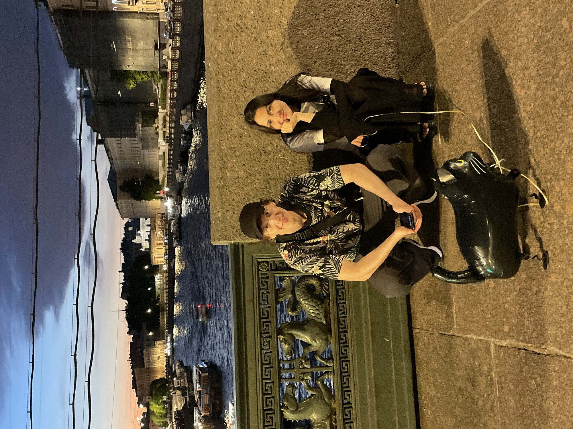 The photograph captures a twilight scene on a bridge in Saint Petersburg, Russia. A man wearing a black baseball cap and a patterned short-sleeved shirt, crouched down, holds a smartphone in his right hand. Beside him, a woman in a light-colored shirt and dark long-sleeved garment, also crouched, rests her chin on her hand, looking towards the camera. In the foreground, a black cat-shaped balloon lies on the cobblestone surface. Behind them, a waterway is visible, with several boats docked and one in motion. The cityscape features buildings illuminated by lights, and the sky displays a gradient of twilight colors with clouds. The bridge itself has a decorative railing with intricate designs.