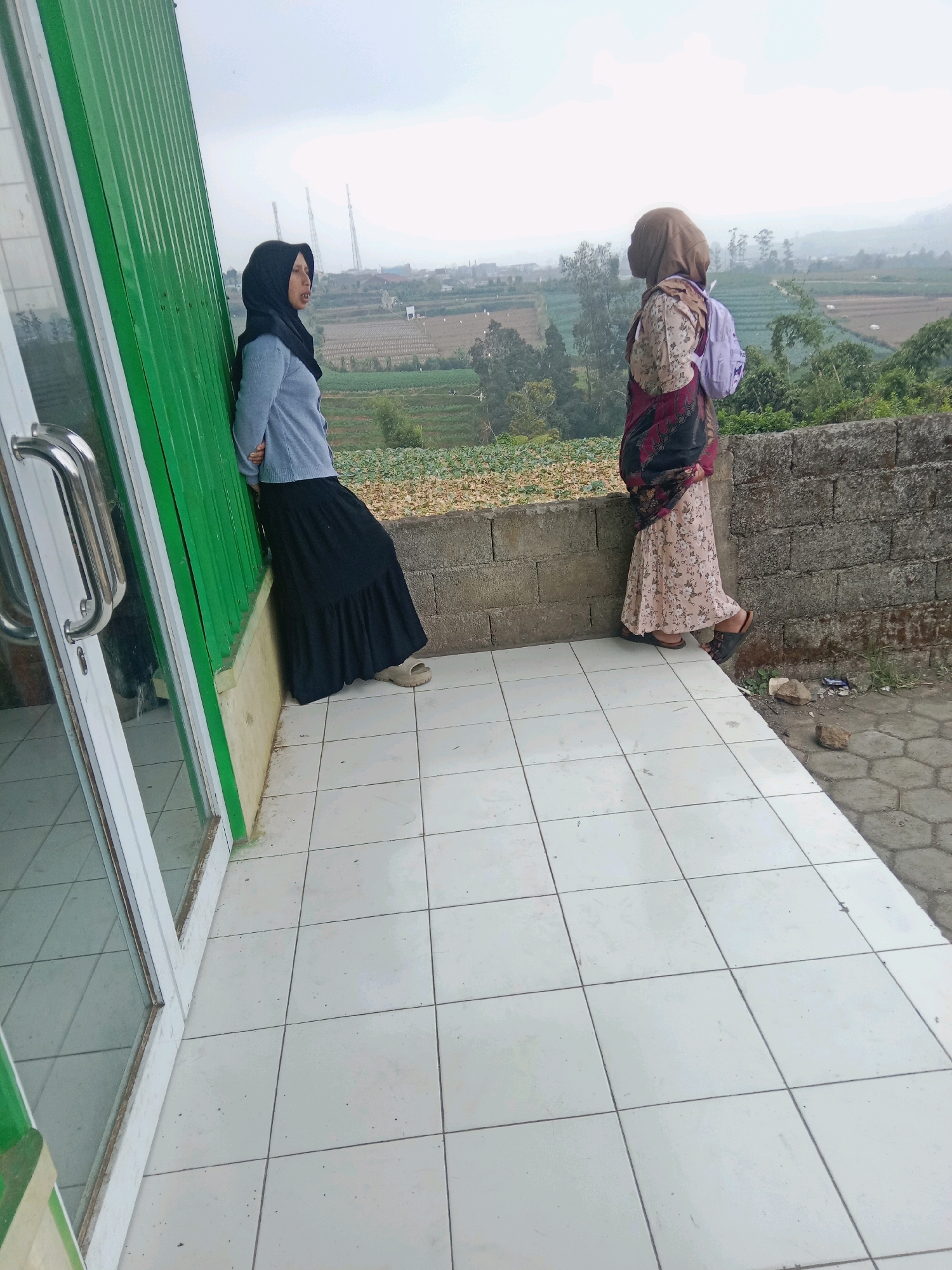 Two women are standing on a tiled porch overlooking a rural landscape in Batur, Indonesia. The woman on the left, wearing a black hijab and a light blue sweater over a black skirt, leans against a green metal wall. The woman on the right, with a brown hijab and a patterned dress, stands facing away from the camera, with a light purple bag on her shoulder. In the background, terraced fields and vegetation are visible under an overcast sky. A low stone wall separates the porch from the elevated landscape. The porch floor is covered with square white tiles, and a portion of a building with a glass door and silver handle is visible on the left.