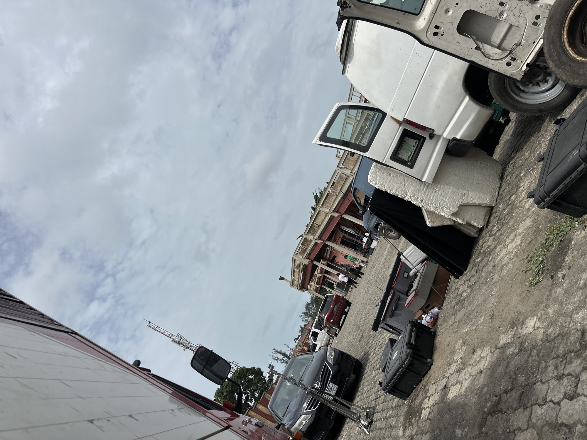 The scene appears to be a cluttered outdoor location, possibly a staging area or parking lot in Lagos, Nigeria. The presence of various vehicles, including vans and cars, suggests a transportation or storage function. There are people visible in the background near a partially constructed or dilapidated building. There are also black cases, some white foam and other loose items scattered on the ground. A communication tower is also visible.