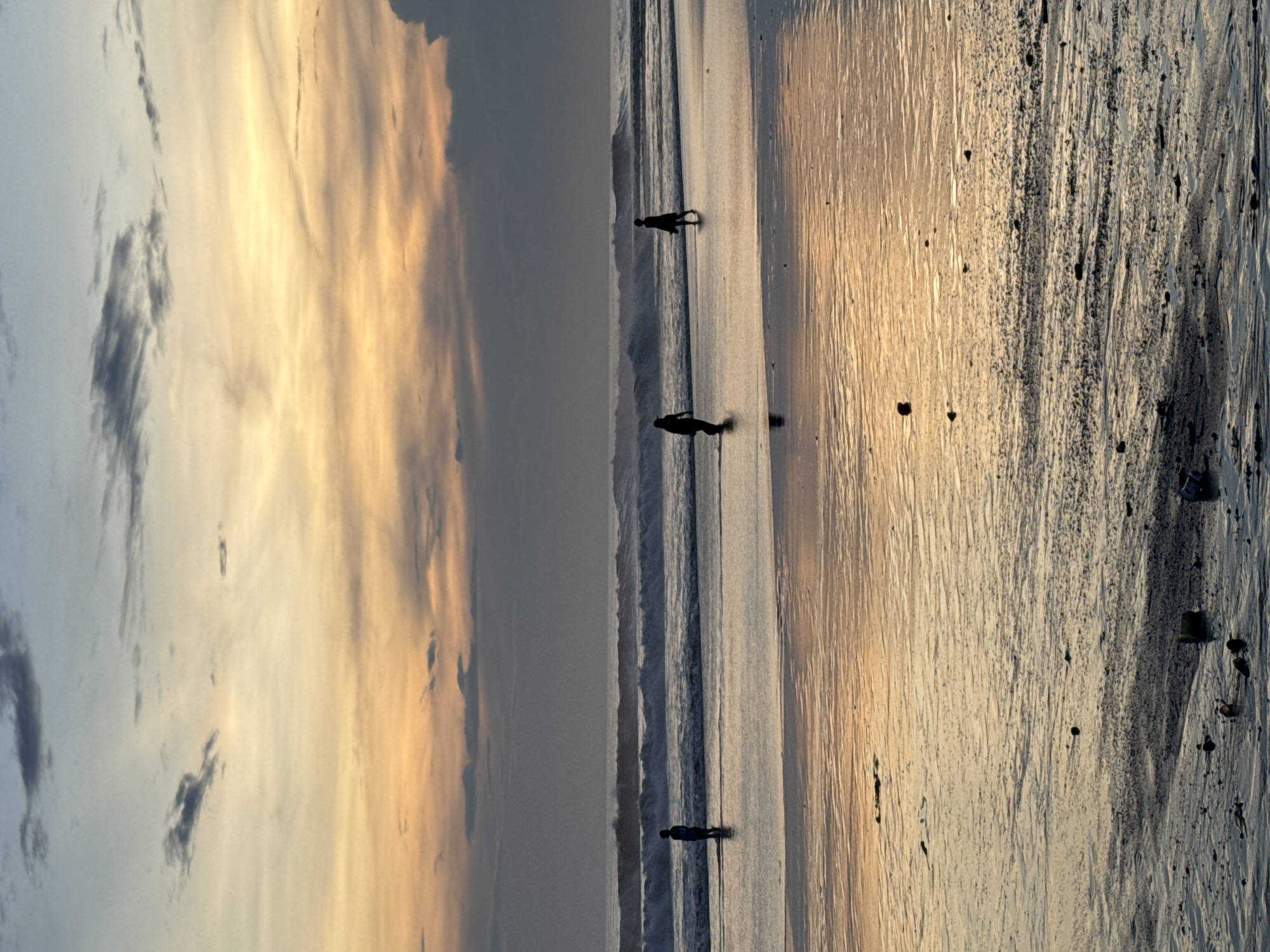 A horizontally oriented image depicts a beach scene in North Kuta, Indonesia, at sunset. The sky is partly cloudy with hues of orange and yellow intermingling with gray and blue. Three people are visible as silhouettes walking along the shoreline. The waves are small and regular. The beach appears wet with some textured sand.