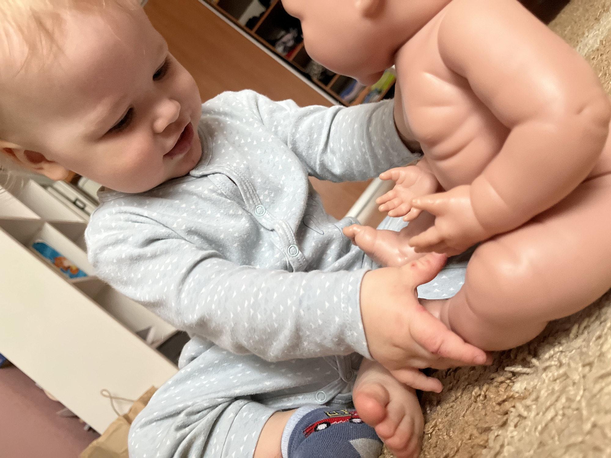 A baby wearing a light blue patterned onesie and a dark blue sock with a red design is holding a nude plastic doll. The baby appears to be touching the doll's foot with one hand and holding the doll with the other. A white shelf unit is visible on the left, and a wooden cabinet is in the background. The scene is on a tan carpet. Location details provided are city of Chaikovsky, Russia.