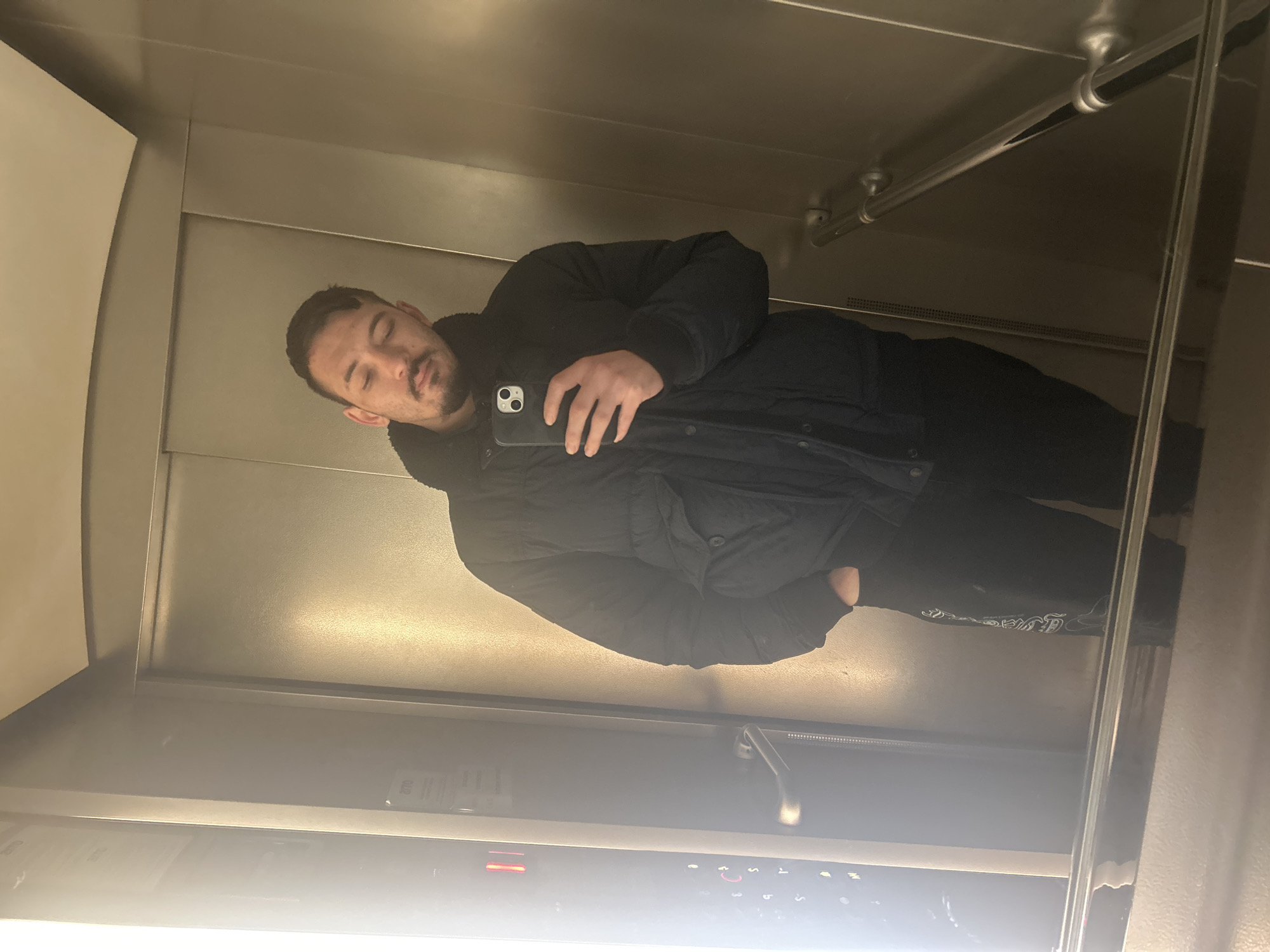 A man is taking a selfie in a mirrored elevator. He is wearing a black puffer jacket and black pants. The elevator has a grab bar and an elevator panel with buttons. The location cannot be determined from the image.