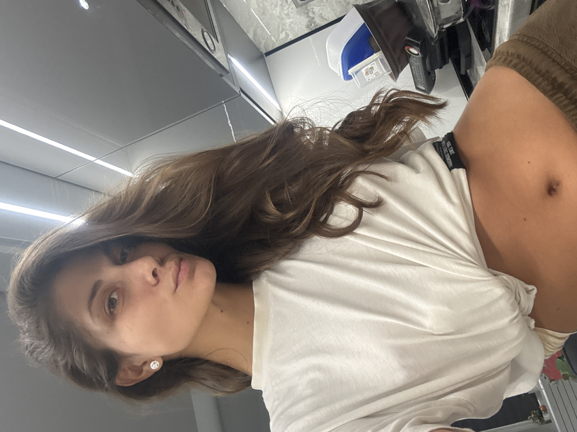 A woman with light brown hair and a white t-shirt poses indoors. The background suggests a modern kitchen, with white cabinets and countertops. The location is undetermined.