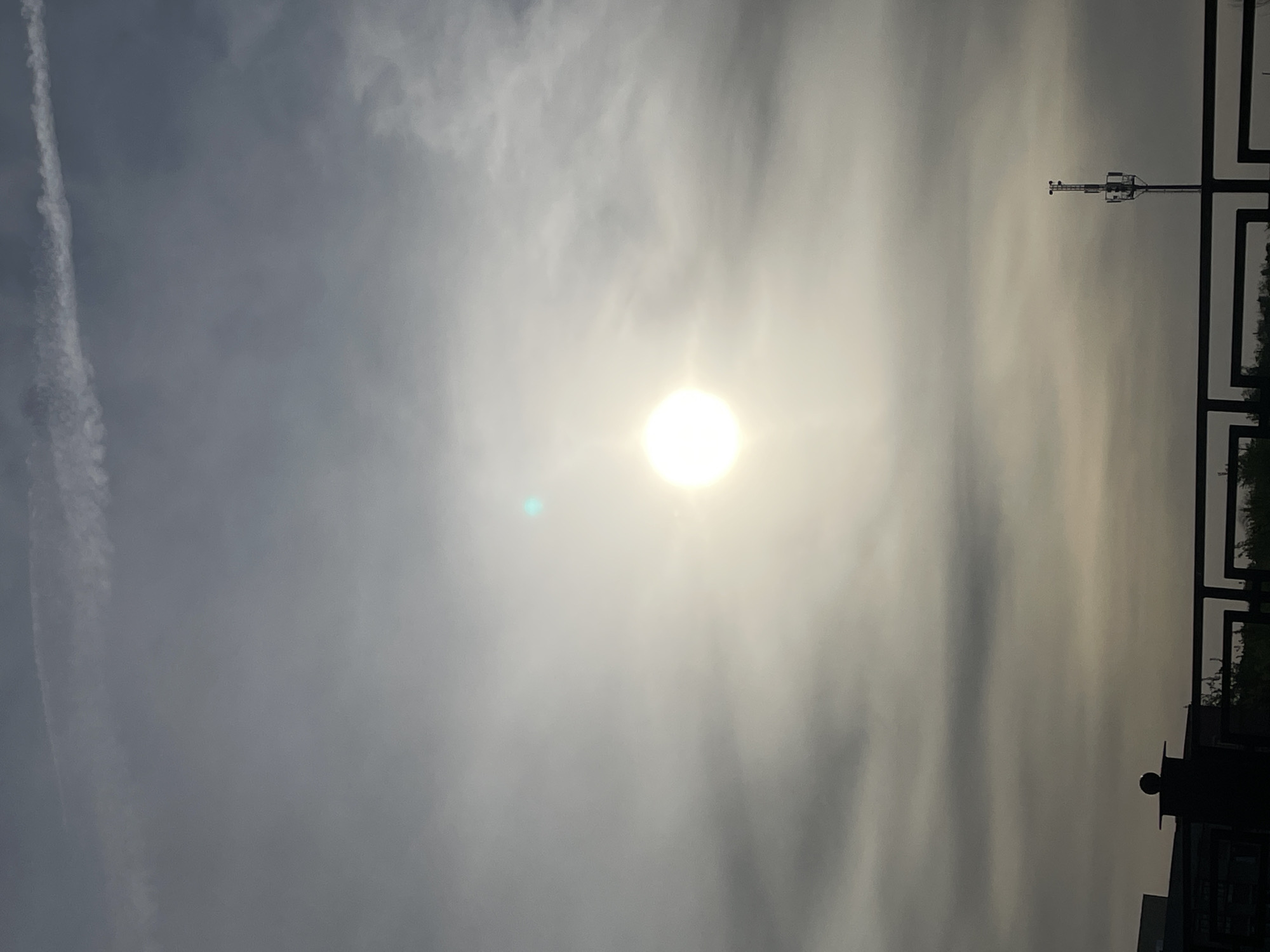The image depicts a bright, overcast sky with the sun prominently visible through the clouds. A contrail is visible on the left side of the image. On the right, there is an object, likely a utility pole, and a dark vertical geometric structure. Based on available evidence, it is not possible to determine the location within Saratov, Russia. There are no identifiable people, subjects, or actions in the image.