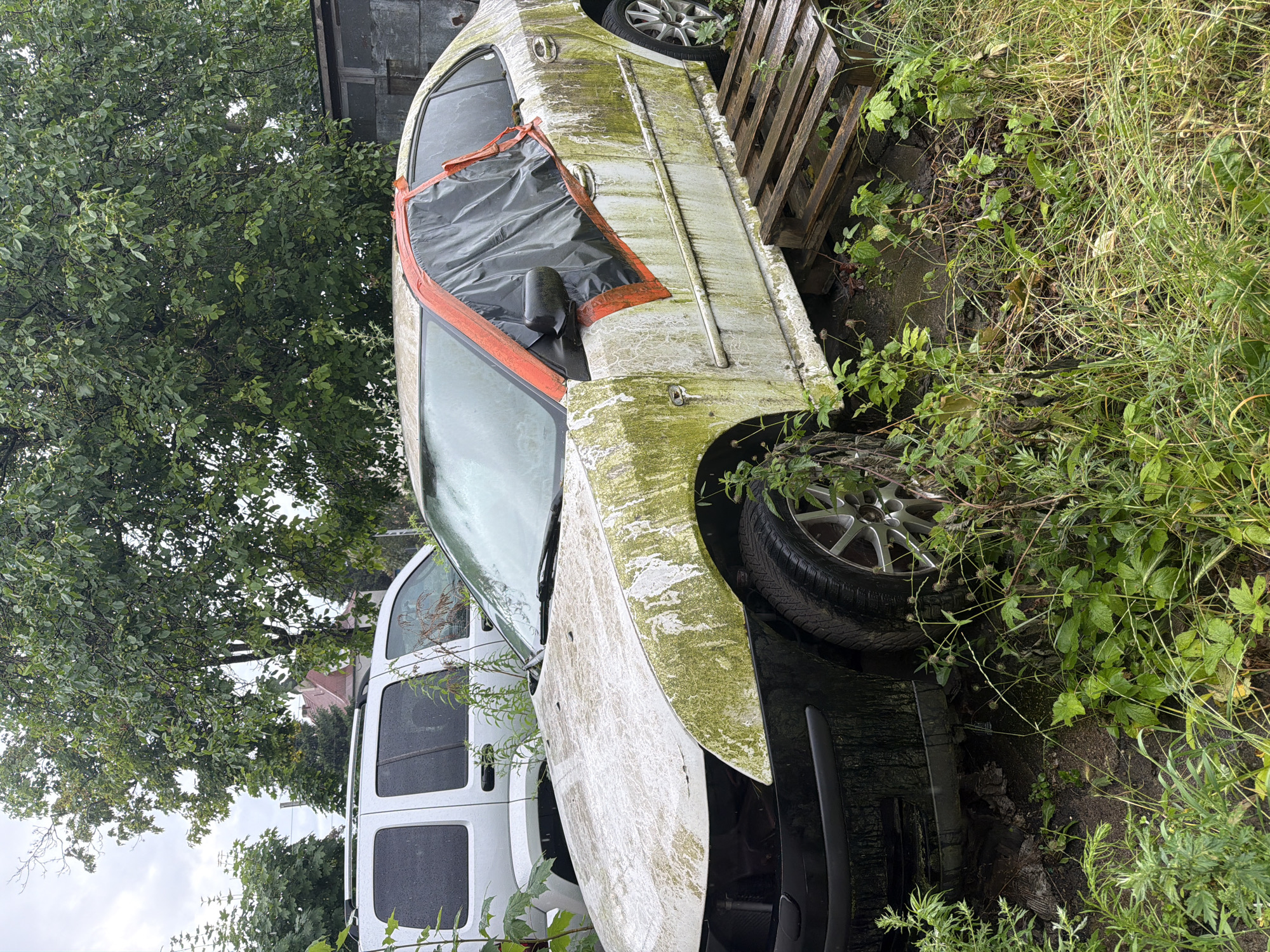The image shows a severely dilapidated and weathered car, likely a sedan, lying on its side amidst overgrown vegetation and what appears to be a scrapyard or abandoned lot. The car's body is covered in patches of green moss or algae, indicating a long period of neglect and exposure to the elements. A large section of the driver's side door is open and covered with a gray tarp secured by bright orange tape, suggesting a makeshift attempt at covering damage or preventing further entry of debris. The car's windows are visible, though their condition is unclear due to the angle and lighting.
In the background, to the left, there's a dense canopy of green leaves from a large tree, with its trunk obscured. Further in the background, a small portion of a building with a red-tiled roof is visible, hinting at a semi-rural or suburban setting. Adjacent to the overturned car, to its right and rear, is a wooden pallet, also appearing weathered and aged.
A portion of a white van is visible in the lower left quadrant of the image, suggesting it's parked nearby or also part of the abandoned collection. The ground beneath and around the car is covered with a mix of mud, debris, and thick, unkempt green grass and weeds, reinforcing the sense of abandonment. The overall impression is one of decay and disuse.
The lighting suggests it might be an overcast day, possibly late afternoon, given the diffused light and the somewhat muted colors. There are no people visible in the image, and the focus is entirely on the state of the abandoned vehicles and their surroundings. The scene evokes a feeling of neglect, perhaps even a touch of melancholy due to the state of the once-functional object now consumed by nature and time. The location context provided is Szczecin, Poland. There is no visible text in the image.