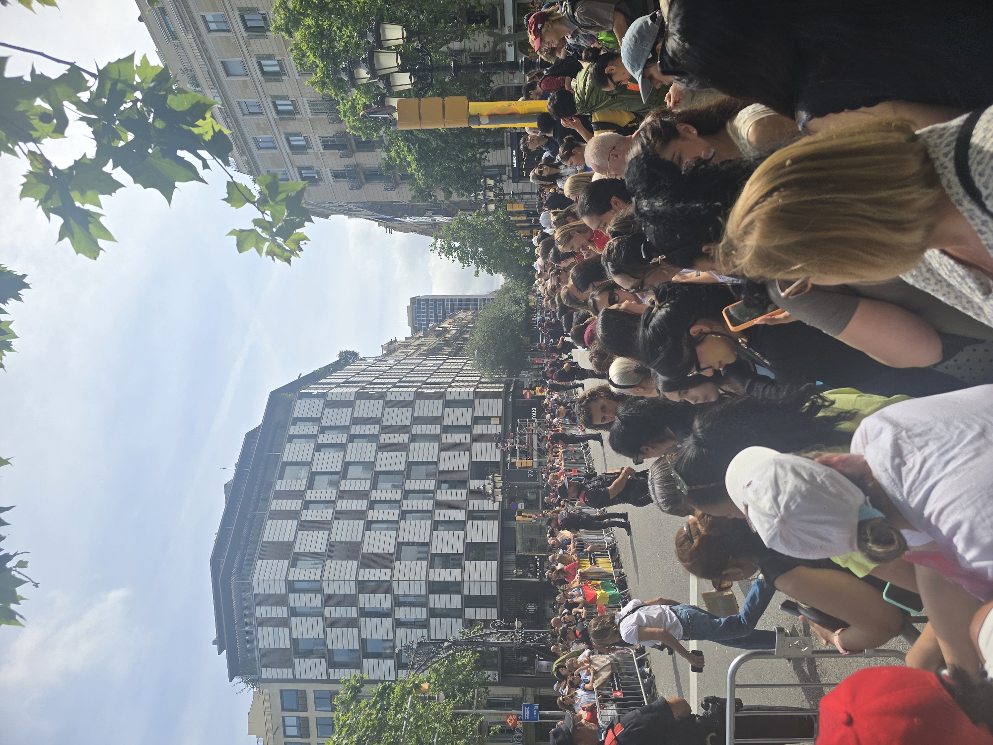 The image depicts a daytime scene in Barcelona, Spain, likely during an event or parade. A large crowd of people are gathered behind metal barricades, suggesting a street event. Several police officers are visible, some in uniform, maintaining order. The background features tall, modern and older buildings, typical of a city environment. Trees line the street, and there are streetlights and traffic signals. Some individuals in the crowd are holding flags, one of which appears to be the flag of Bolivia. The people are diverse in age and gender, and many are looking towards something outside the frame, indicating they are observing an event. The overall atmosphere appears to be one of public gathering and observation.