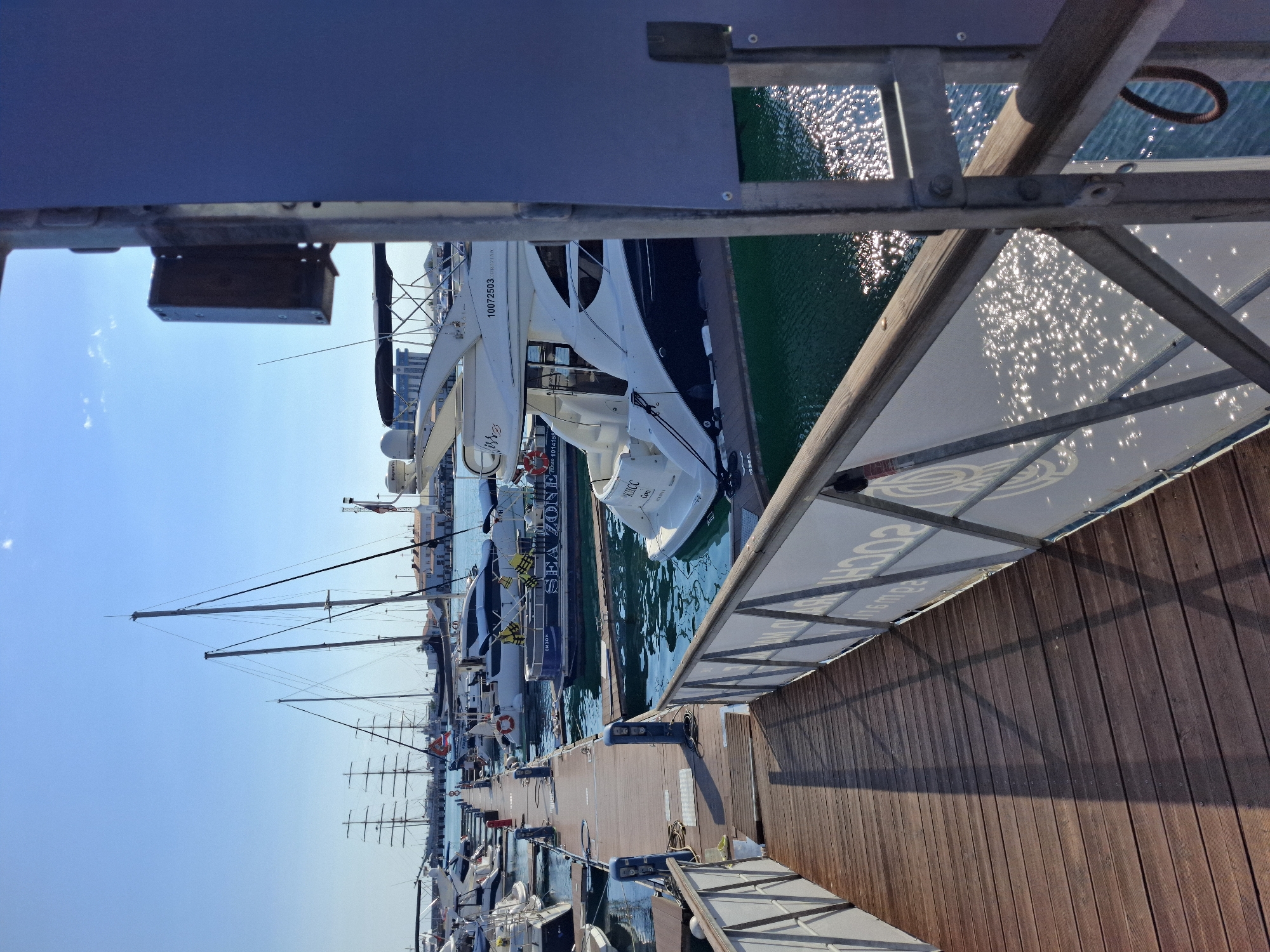 This media file depicts a view of a marina in Sochi, Russia. The scene is dominated by several yachts and sailboats moored along a wooden pier. The water is a vibrant green, reflecting the bright sunlight which creates a shimmering effect on the surface. The sky is a clear, bright blue, suggesting a sunny day. The orientation of the image is such that the bottom of the frame points upwards towards the sky and the top of the frame points downwards towards the water.
The foreground features a wooden pier with metal railings. A white boat with a dark hull is docked on the left, and the masts of several sailboats extend upwards towards the sky. In the mid-ground, more yachts are visible, including a prominent white yacht with the text "10072503 SOCHI" and "MRS" visible on its side. Another yacht has the word "SEA" in large letters. The background shows a cityscape with buildings.
The time of day appears to be daytime, likely late morning or early afternoon, given the strong sunlight and clear sky. The weather is pleasant and sunny. The overall impression is one of a well-maintained and active marina on a beautiful day in Sochi. There are no people clearly visible in the image, focusing instead on the vessels and the marina infrastructure.