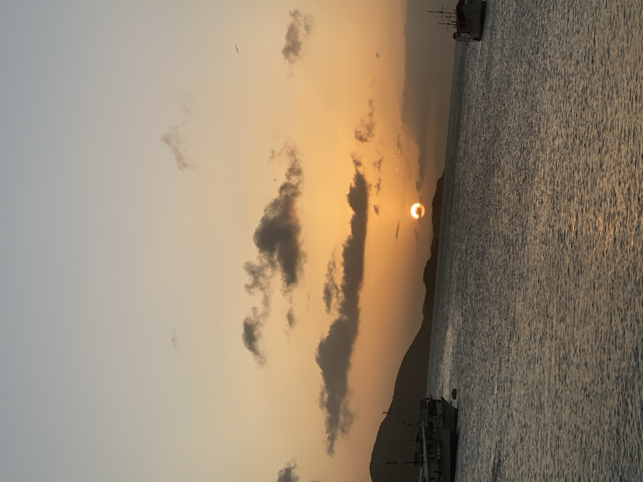 An idyllic sunset scene captures the sun partially obscured by dark, vertical clouds over a body of water. A vague mountain silhouette is discernible under the sun. The water reflects the golden and orange light. A vessel is visible in the upper right corner and a boat or structure in the lower right corner of the frame. The sky transitions from a light gray to a warm orange near the horizon. A few birds are visible in the sky.