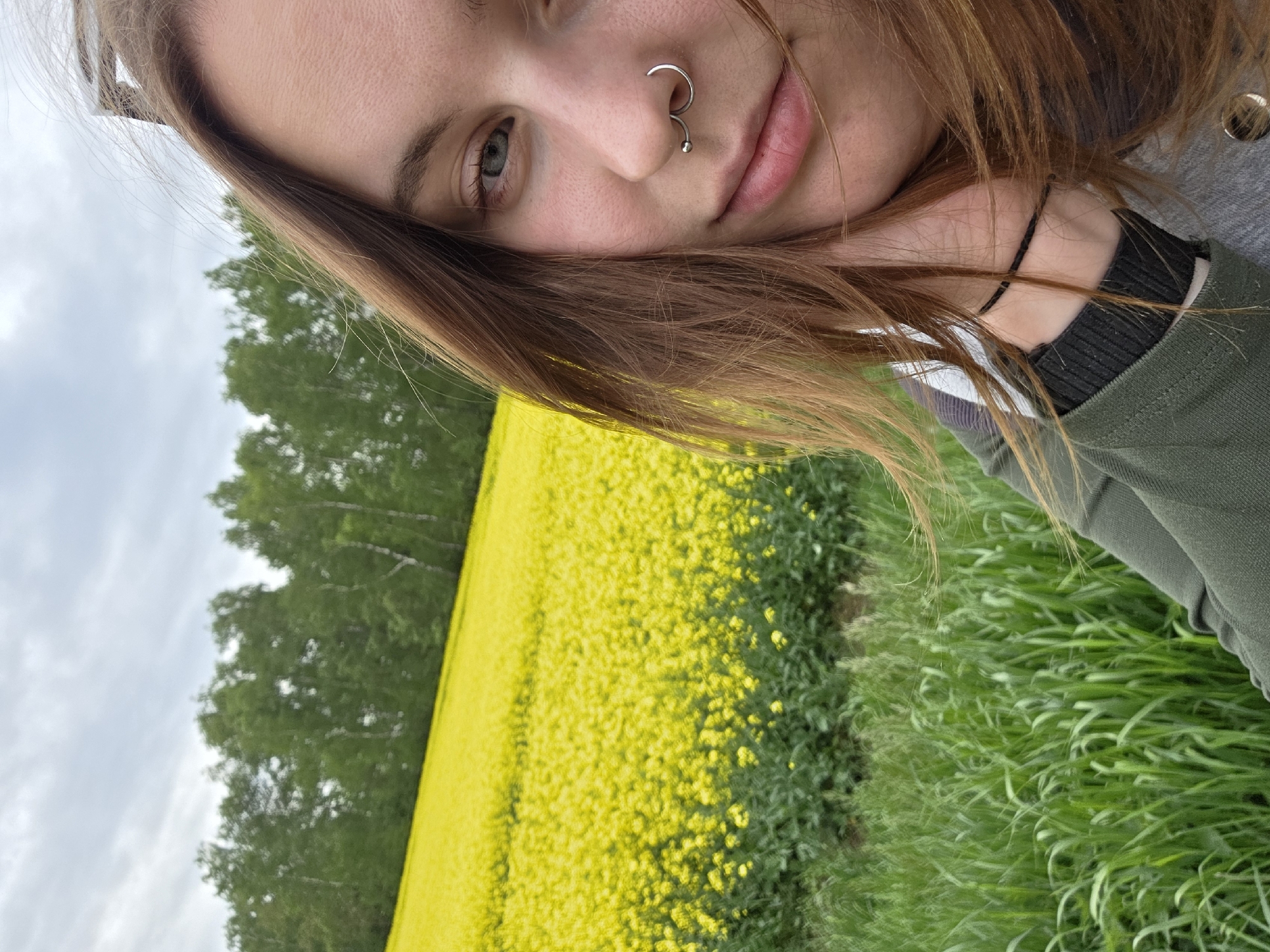 A selfie of a young woman with light brown hair and a nose ring is taken outdoors with a yellow field of flowers and green trees in the background under a cloudy sky. The woman is wearing a green and grey jacket. Her face is partially visible, and she is resting her chin on her hand. The photograph appears to be taken at a slight angle. The location is unknown.