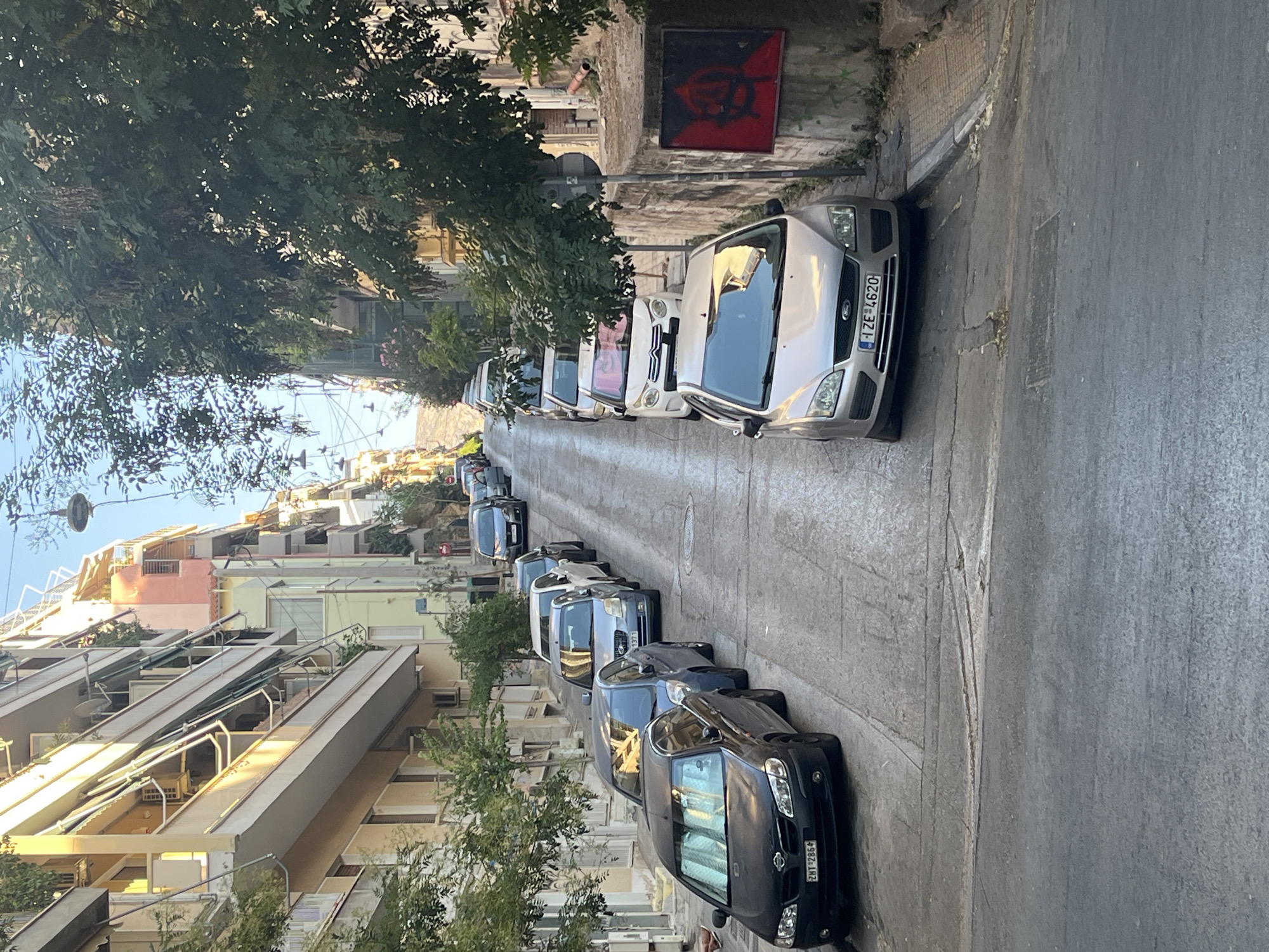 A narrow, steep street in Athens, Greece, is lined with parked cars and apartment buildings. The buildings, in shades of cream, yellow, and pink, feature balconies with awnings. Trees with lush green foliage are interspersed along the street. Several cars are parked on both sides of the road, suggesting a residential area. On the right side of the street, a red square with a black symbol, possibly an anarchist symbol, is painted on a concrete wall. The overall impression is of a typical urban street scene in Athens on a sunny day.
