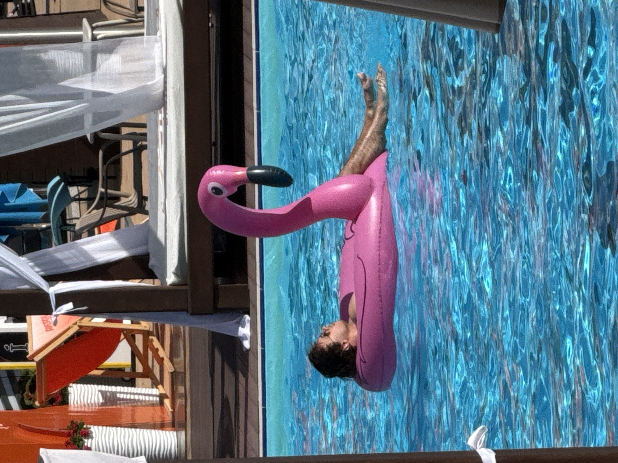 In Khimki, Russia, a young man with short, dark hair and sunglasses is relaxing on a pink flamingo inflatable in a bright blue swimming pool. He is lying on his back, with his head propped up on the edge of the inflatable. His legs are extended and submerged in the water, and his feet are visible. The flamingo inflatable is positioned in the center of the pool, with its head and neck arching upwards. The pool water is clear and reflects the bright sunlight. In the background, a wooden deck surrounds the pool, with lounge chairs and umbrellas visible. The scene appears to be a resort or a private residence.