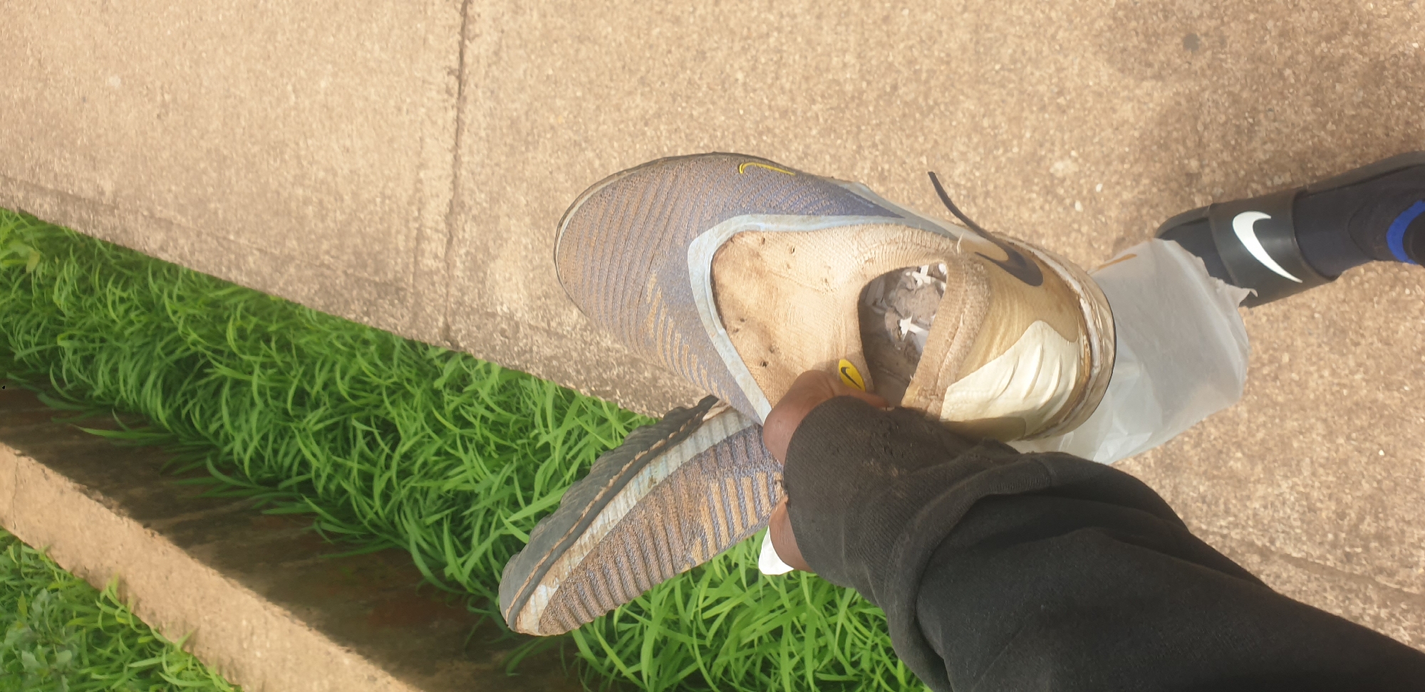 In Barakin Lerawi, Nigeria, a person's hand, clad in a black long-sleeved garment, holds a dirty, tan and blue Nike athletic shoe. The shoe appears to be a slip-on style with a woven upper, showing significant wear and tear, and is filled with debris. The shoe is being held over a white plastic bag, which is partially visible. The person is also wearing a black Nike slide sandal over a dark sock with blue accents on their visible foot. The background consists of a concrete pathway on the right and a concrete drainage ditch filled with lush green grass on the left.