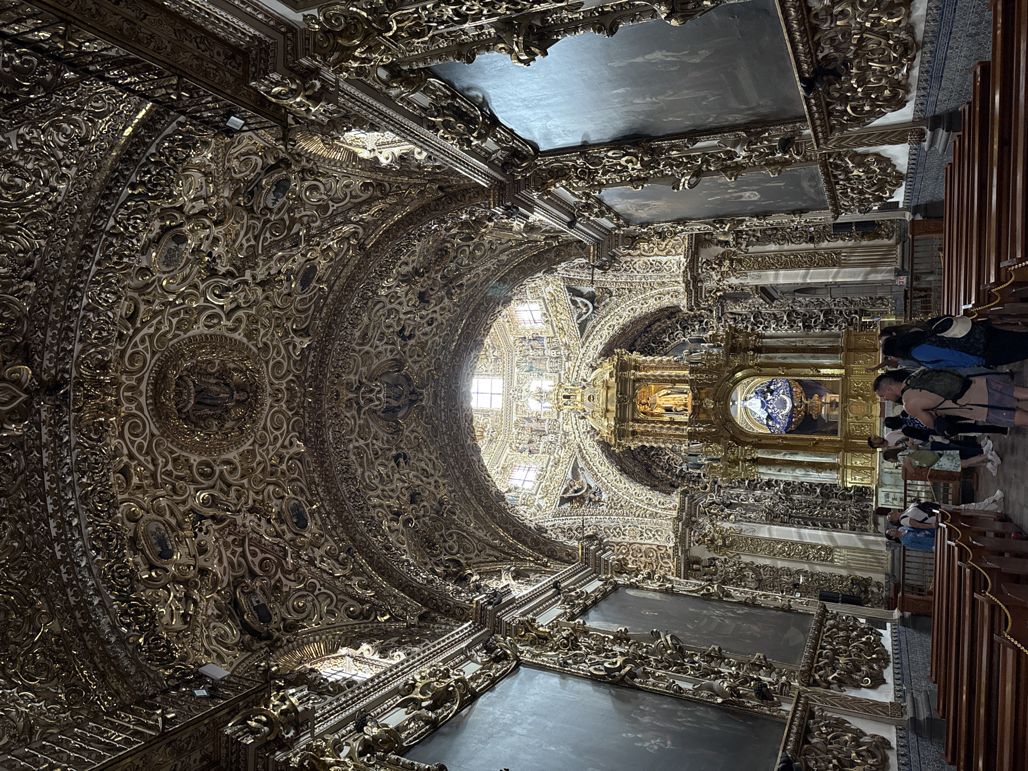 The image captures the exceptionally ornate interior of a church or chapel, characterized by an overwhelming display of gilded Baroque or Churrigueresque architecture. This style, particularly the extensive use of gold leaf and intricate carvings, strongly suggests a location like the Capilla del Rosario in Puebla, Mexico, which is renowned for its elaborate decoration.
The vaulted ceiling and arches are completely covered in gold leaf, featuring incredibly detailed scrollwork, floral patterns, angelic figures, and other architectural embellishments. Numerous framed paintings and frescoes are integrated into the gilded surfaces, depicting figures and scenes, some against sky-like backdrops. Natural light filters in through upper windows, complemented by artificial light fixtures visible throughout the elaborate ceiling.
Towards the center of the image, a magnificent golden altarpiece or reredos stands as a focal point, adorned with statues, columns, and intricate gold carvings, including a central figure that appears to be robed in blue.
On the right side of the interior, traditional wooden pews are arranged, accommodating several visitors. These individuals, dressed in casual attire, appear to be tourists or sightseers, standing and seated, observing the lavish decorations and absorbing the grandeur of the space. The lower right wall or floor section shows distinct blue and white patterned tiles, a characteristic element often associated with Talavera pottery from Puebla.
The scene conveys a sense of awe and reverence, typical of visitors encountering such a historically and artistically significant religious edifice. The overall impression is one of immense detail and opulence, captured during daylight hours. No discernible text is visible within the image.