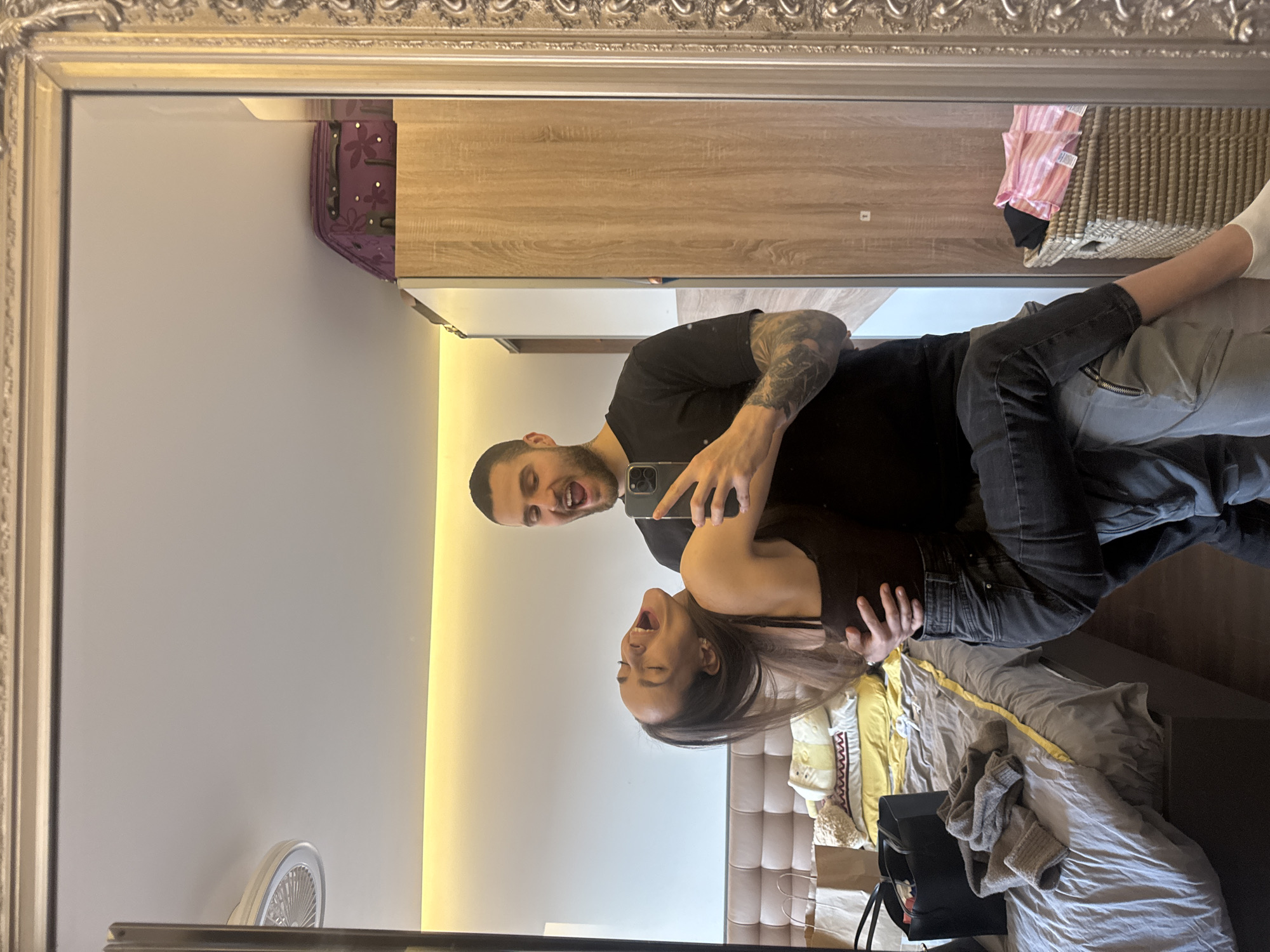 The image captures a couple taking a selfie in a large ornate mirror. They are likely in a bedroom. The man has a visible arm tattoo and is holding a smartphone. The woman is leaning into him, both are laughing. Behind them, the reflection shows a bed with light grey bedding, a headboard, a light wood cabinet or wardrobe, and other miscellaneous items. It's impossible to determine the exact location in Sofia, Bulgaria, solely from the image.