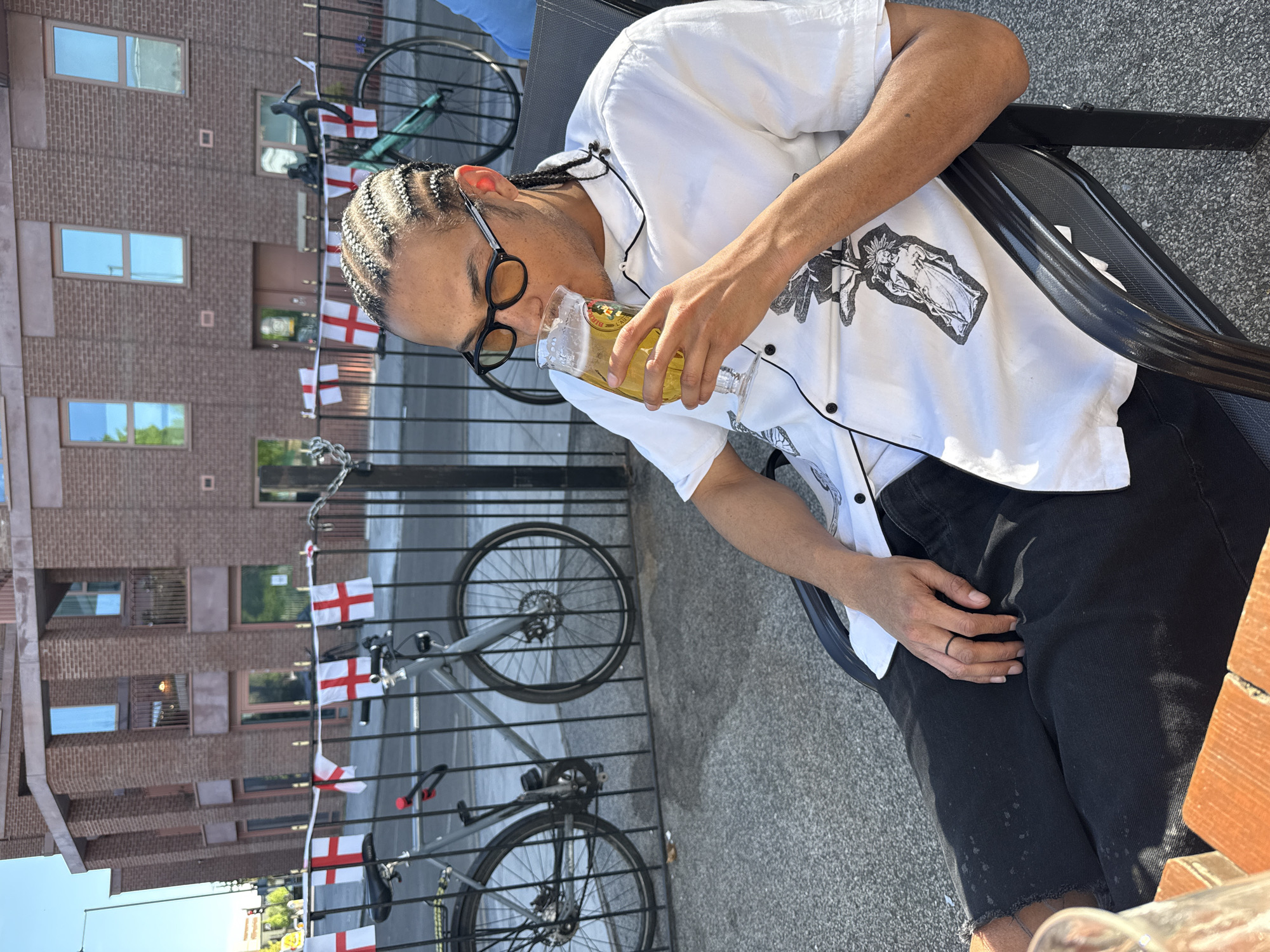 A person with braided hair and glasses is sitting outdoors, drinking from a glass of yellow liquid. The person is wearing a white short-sleeved button-down shirt with a black and white graphic design and black shorts. The person is seated on a black metal chair. A metal railing with the flag of England is behind the person, with two bicycles parked next to it. A brick building is visible in the background, suggesting an urban location. The scene appears to be at an outdoor pub or restaurant. Given the English flags, the location is likely in London, United Kingdom.