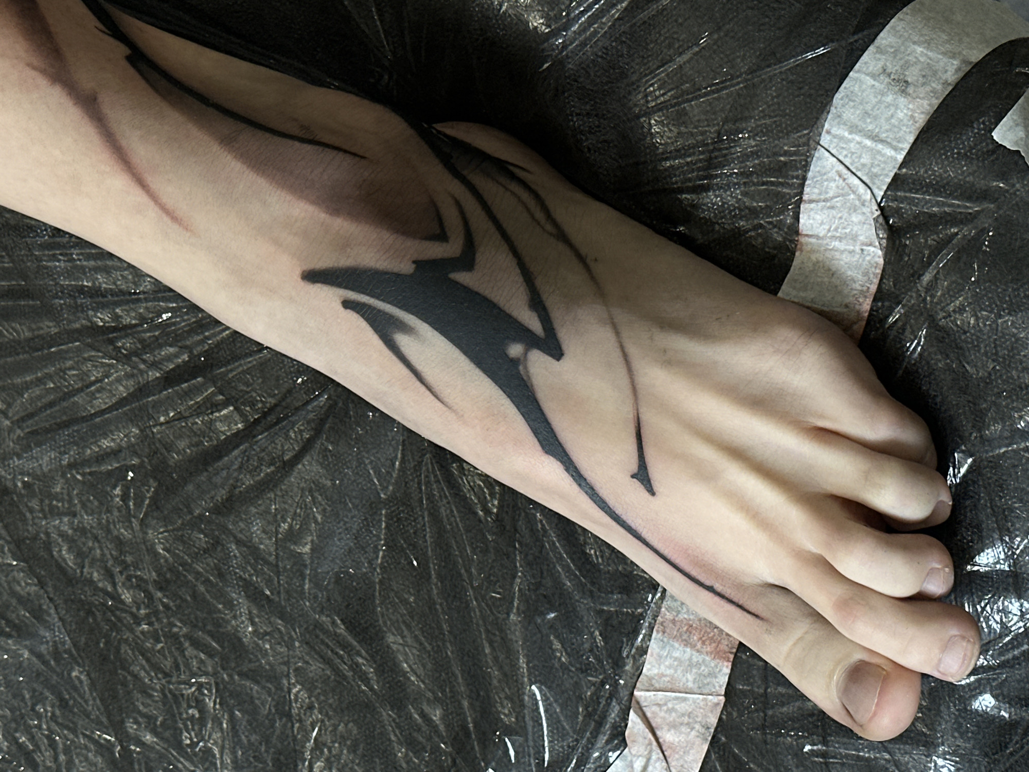 The image displays a close-up view of a person's right foot and lower leg, featuring a newly applied black tattoo. The limb rests upon a dark, crumpled plastic sheet, likely used for protection during a tattooing process.
The tattoo, executed in black ink, is an abstract, dynamic design that covers the top of the foot and extends upwards onto the ankle. It comprises a central, solid black, angular shape resembling a stylized bolt or a sharp, organic form. From this core design, thinner, delicate lines and subtle shading radiate across the skin, creating a sense of movement or a wispy effect. The aesthetic is contemporary, possibly drawing from tribal or graphic design influences. The fair skin tone of the individual provides a strong contrast to the dark ink, highlighting the tattoo's details. Some areas around the tattoo exhibit slight redness, suggesting the work is fresh.
The toes are visible towards the right of the frame, showing natural, unpolished toenails. The background is dominated by the dark, textured plastic sheeting, which reflects light in various spots, creating subtle highlights. A strip of white adhesive tape or paper, bearing faint reddish-brown marks (possibly ink or residue), is visible on the plastic surface towards the right. The overall setting implies an indoor environment, such as a tattoo studio in Moscow, Russia. No visible text is present in the image.