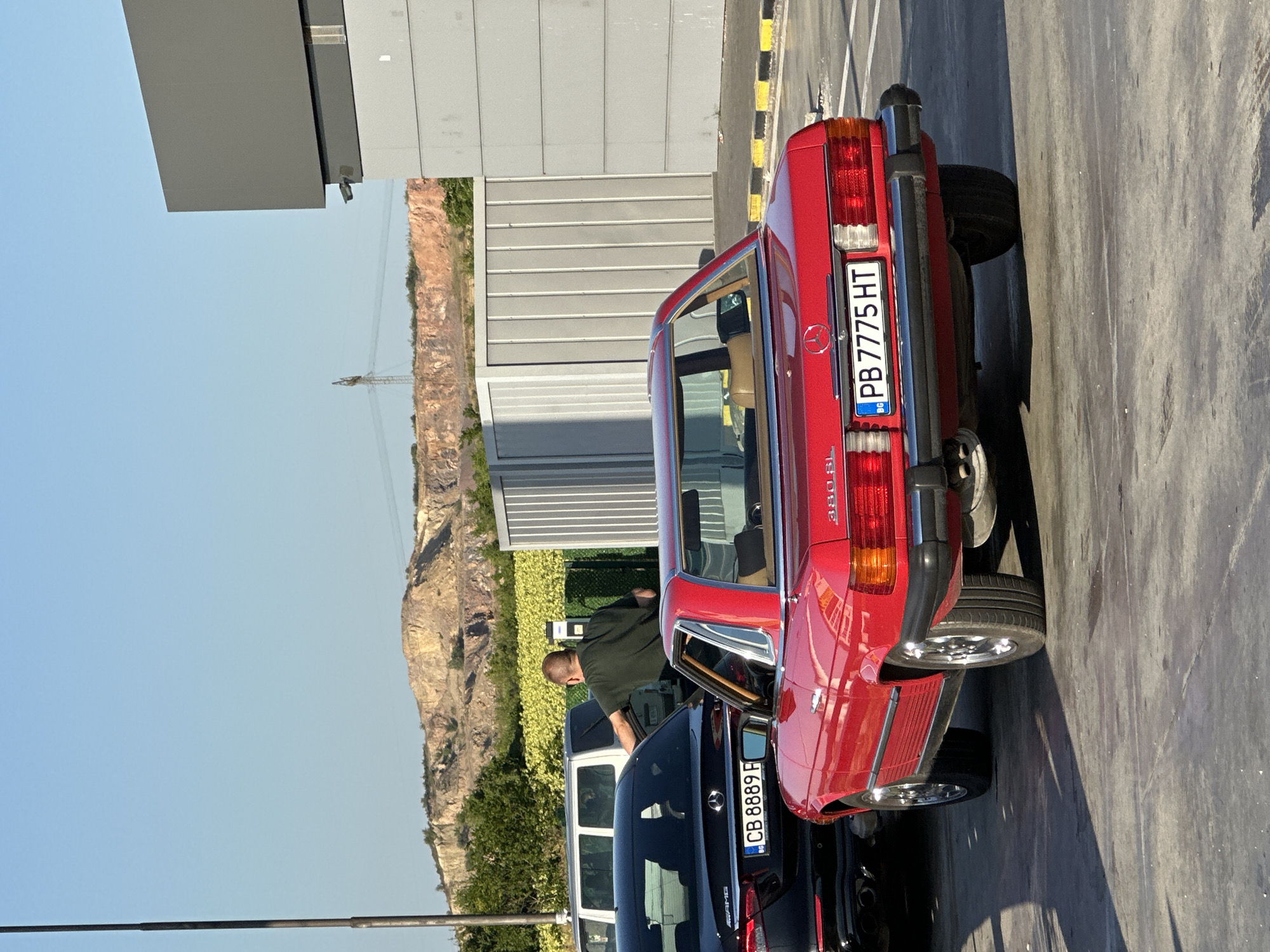 The image captures a sunlit outdoor scene, likely in the vicinity of Polsko Padarevo, Bulgaria, with a quarry or excavated hillside visible in the background under a clear blue sky. In the foreground, a bright red classic Mercedes-Benz SL model is parked, its rear facing the camera. The car displays a Bulgarian license plate "PB 7775 HT". To the left of the red car, the rear of a black Mercedes-Benz sedan is partially visible, with a Bulgarian license plate "CB 8889 F". A man in a dark green t-shirt is seen standing between the two vehicles, with his right arm extended towards the black sedan, possibly interacting with its driver or passenger. A white van is parked behind the black sedan. In the background, a large grey industrial-style building or structure with ribbed panels stands to the right, adjacent to a lighter grey facade. A utility pole and wires are also visible against the backdrop of the quarry. The ground appears to be asphalt or a similar paved surface, with painted yellow and black lines marking the edge of a parking area.