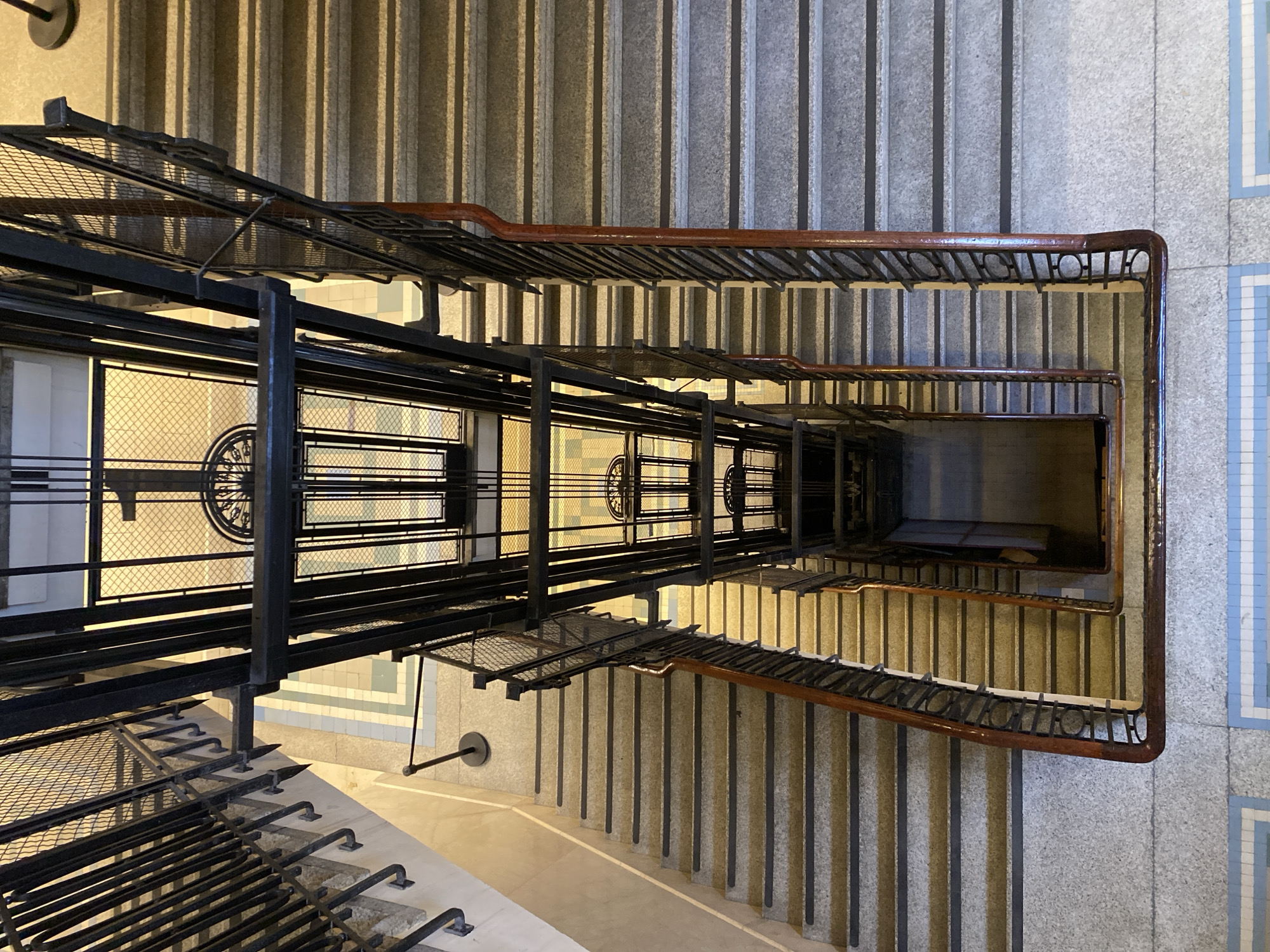 An interior shot from above looking down a staircase with an antique elevator in the Casa de Gobierno in Buenos Aires, Argentina. The elevator has a black metal frame and decorative details. The staircase is lined with gray stone and has a wooden handrail. The floor is tiled in a light blue and white pattern. The overall lighting is soft and diffused. No people are visible.