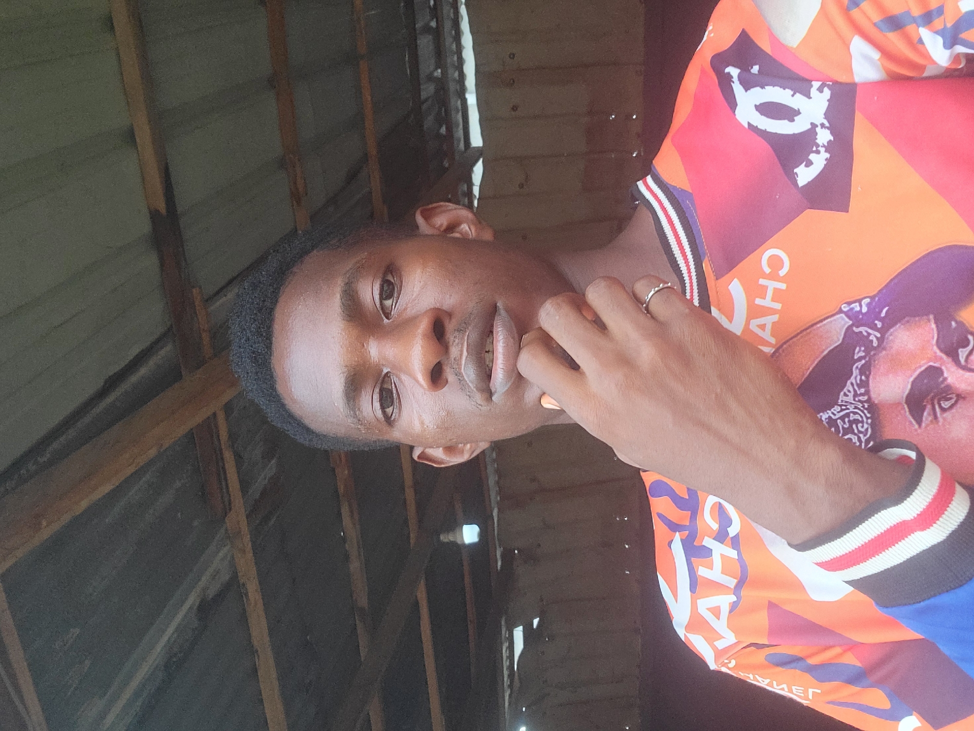 In Egbagu, Nigeria, a young Black man is captured in a medium shot, standing in front of a weathered, open-air structure with a corrugated metal roof supported by wooden beams. He is wearing a vibrant, multi-colored shirt featuring graphic designs, including what appears to be the Chanel logo and a portrait of Tupac Shakur. His right hand is raised to his chin, with a ring on his index finger. The background consists of a rustic, dimly lit enclosure, possibly a workshop or storage area, with corrugated metal walls. A single bare lightbulb hangs from the ceiling, providing illumination.