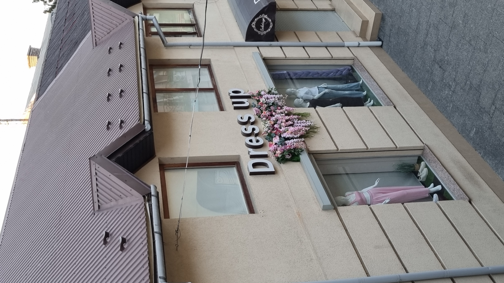 The image shows the facade of a building, likely a retail establishment, in Mukachevo, Ukraine. The building has a light-colored, textured exterior and features large display windows. Above the windows, a sign with the words "Dress up" is mounted. The sign is accompanied by a decorative arrangement of artificial flowers in shades of pink and purple. In the display windows, mannequins are visible, one dressed in a pink two-piece outfit and another pair dressed in casual attire. The building has a dark brown, corrugated metal roof with several dormer vents. Adjacent to the "Dress up" sign, a dark awning with a circular logo is visible, possibly indicating another business. The surrounding area appears to be paved with gray stone tiles. The overall scene depicts a commercial street in a European city.