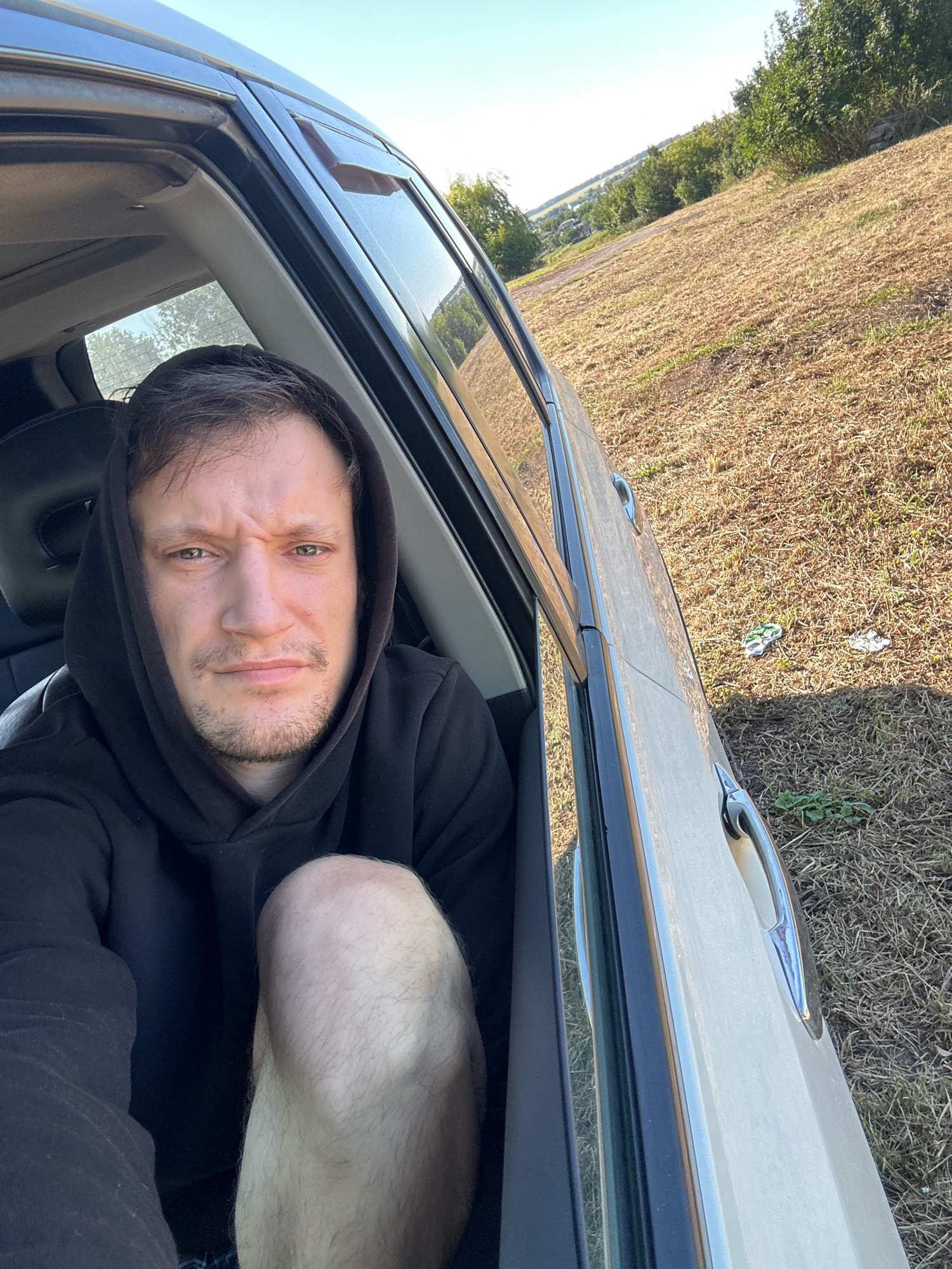 A man wearing a black hoodie is seated in the driver's side of a light-colored vehicle, with the door open and the camera positioned inside, facing forward. The man has short, dark hair and is looking directly at the camera with a serious expression. His right knee is visible, with light hair on the leg. Outside the vehicle, a dry, grassy field stretches towards a line of trees and a hazy sky. Some litter is visible on the ground near the car door. The scene appears to be located in a rural or suburban area, possibly on the outskirts of Makiv, Ukraine, given the landscape and vegetation. The lighting suggests it is daytime, likely late afternoon due to the angle of the sun creating shadows.