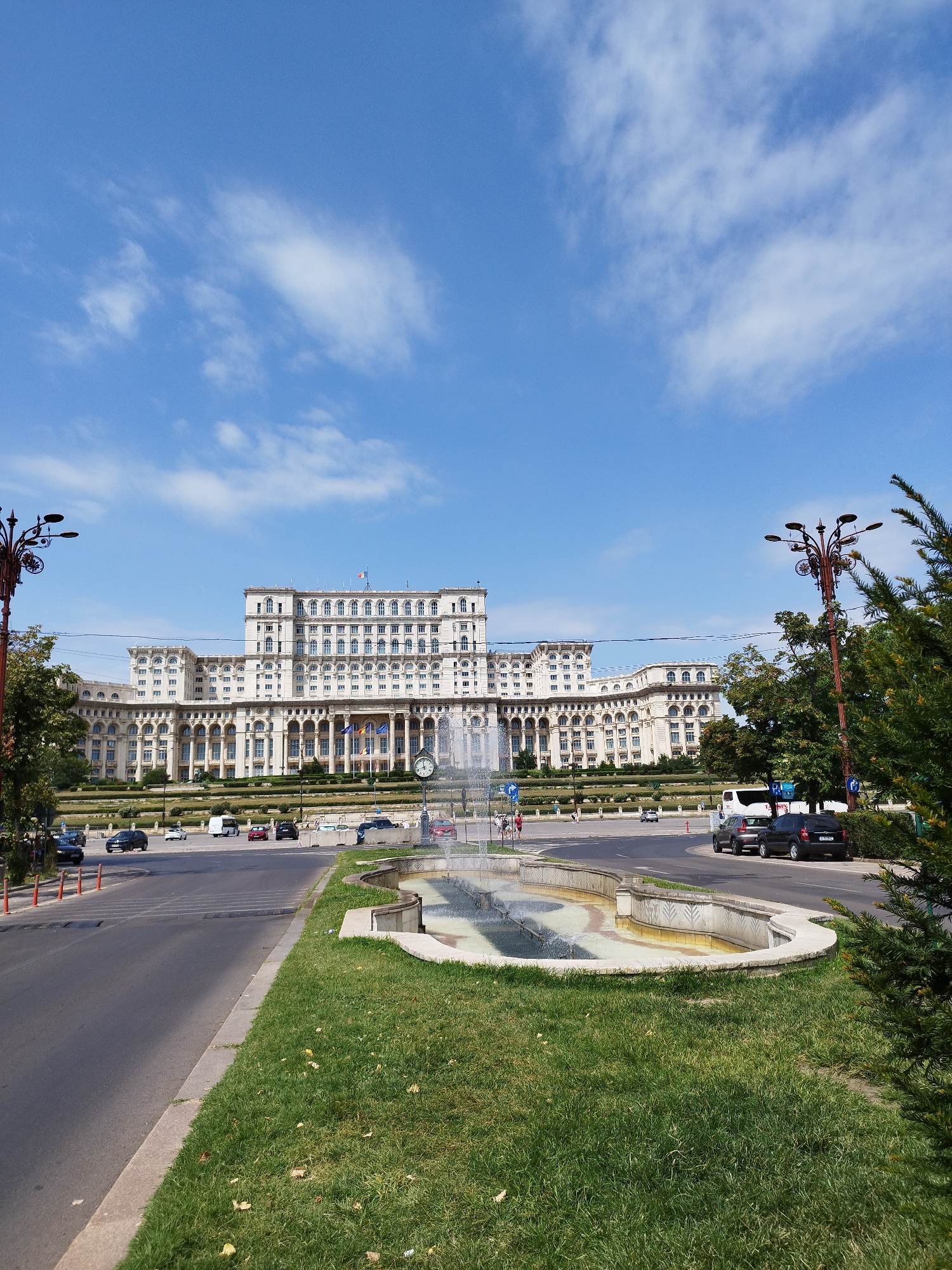 The image shows the Palace of the Parliament, a monumental building in Bucharest, Romania, on a clear day with a blue sky and scattered clouds. In the foreground, a street curves around a grassy area with a decorative fountain. Cars are driving on the street and parked on the side. A lamppost with an ornate design stands on the left side of the image. The Palace of the Parliament dominates the background, displaying its grand architecture with numerous windows and columns. The scene depicts a busy urban environment with a mix of traffic and pedestrian activity.