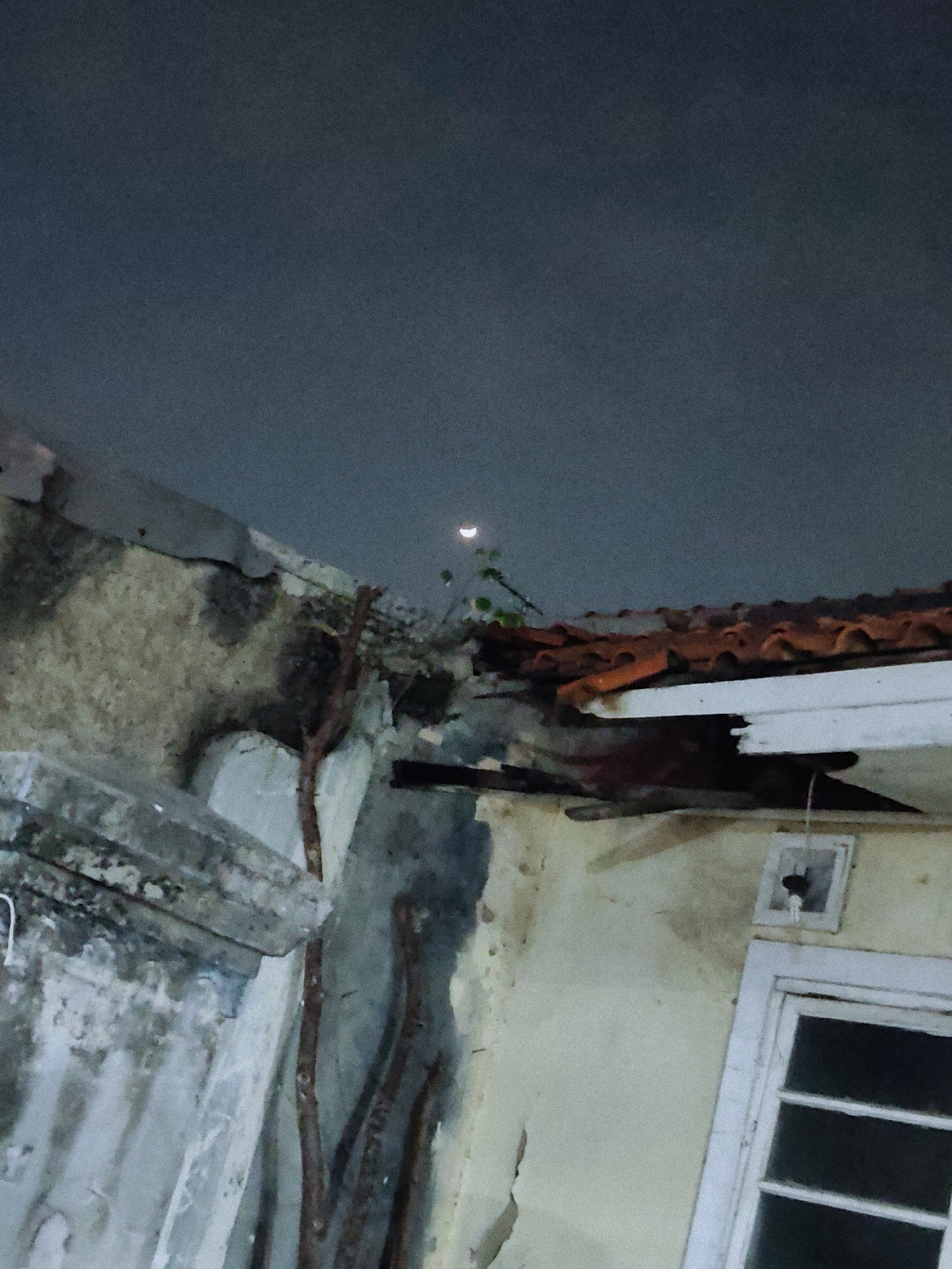 In the city of Mekarmukti, Indonesia, an exterior shot captures a night sky with a sliver of a crescent moon visible. The foreground is dominated by the dilapidated structure of a building, featuring a broken tiled roof. Some sections of the building's wall appear damaged and discolored, with exposed rebar suggesting structural decay. Green foliage is seen growing near the roofline. A window frame with a partially obscured glass pane is visible on the right side of the frame. The overall impression is one of neglect and disrepair.