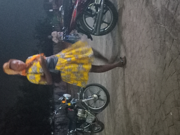 A woman in a yellow dress walks near parked motorcycles. She is wearing a headscarf and an orange shawl. The motorcycles have shiny wheels. The ground is uneven and textured. The background is dark, possibly indicating nighttime. Other people are vaguely visible in the distance. The woman is captured mid-stride, with her dress slightly flared.