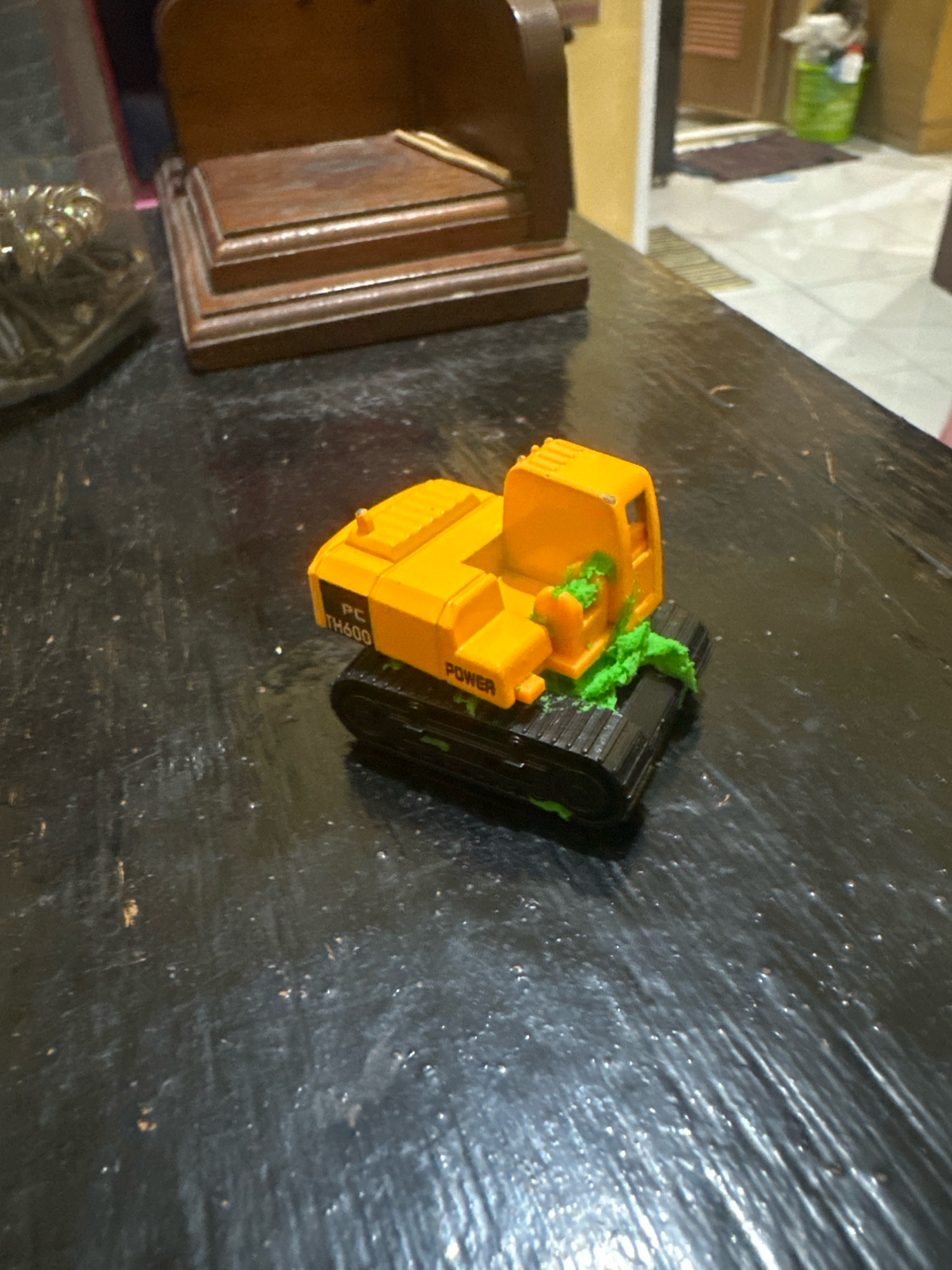 An orange and black toy excavator, with some green material smeared on its tracks, sits on a dark, textured surface. The excavator has the text "PC TH600" and "POWER" visible on its side. In the background, a wooden tiered object is visible, along with other objects and a doorway leading to a tiled floor and a green trash can. The scene appears to be indoors, potentially in a home or workshop environment. No specific location in the city of Saruni, Indonesia, can be determined from the image.