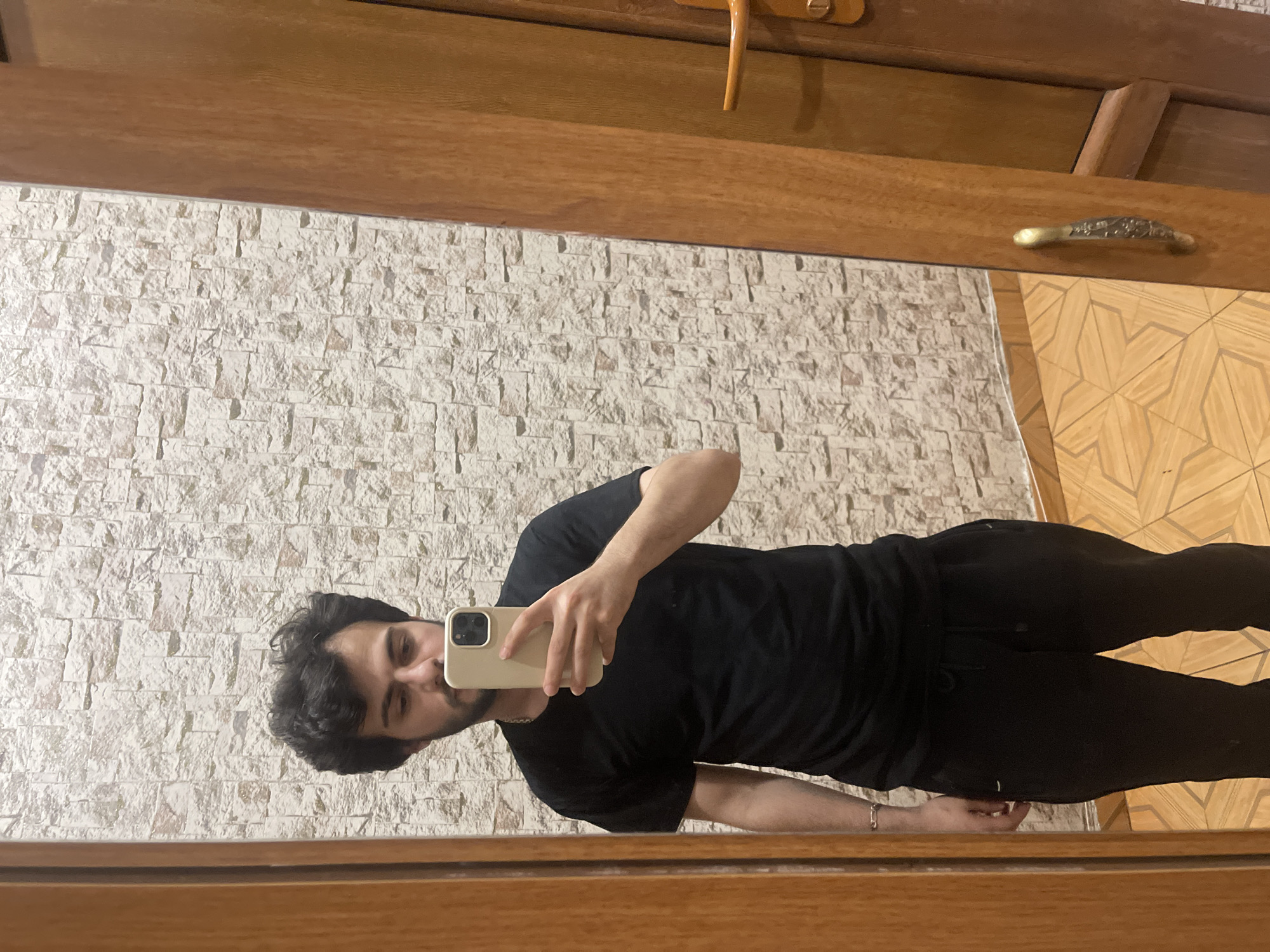 A young man with dark, curly hair and a beard takes a mirror selfie in a room. He is wearing a black t-shirt and black sweatpants, and a silver bracelet is visible on his left wrist. He is holding a beige smartphone in his right hand. The background consists of a textured, light-colored stone wall and a wooden door with a brass handle. The floor has a geometric pattern in shades of brown and yellow. The scene appears to be inside a home or accommodation, possibly in Tbilisi, Georgia, given the architectural style and materials.