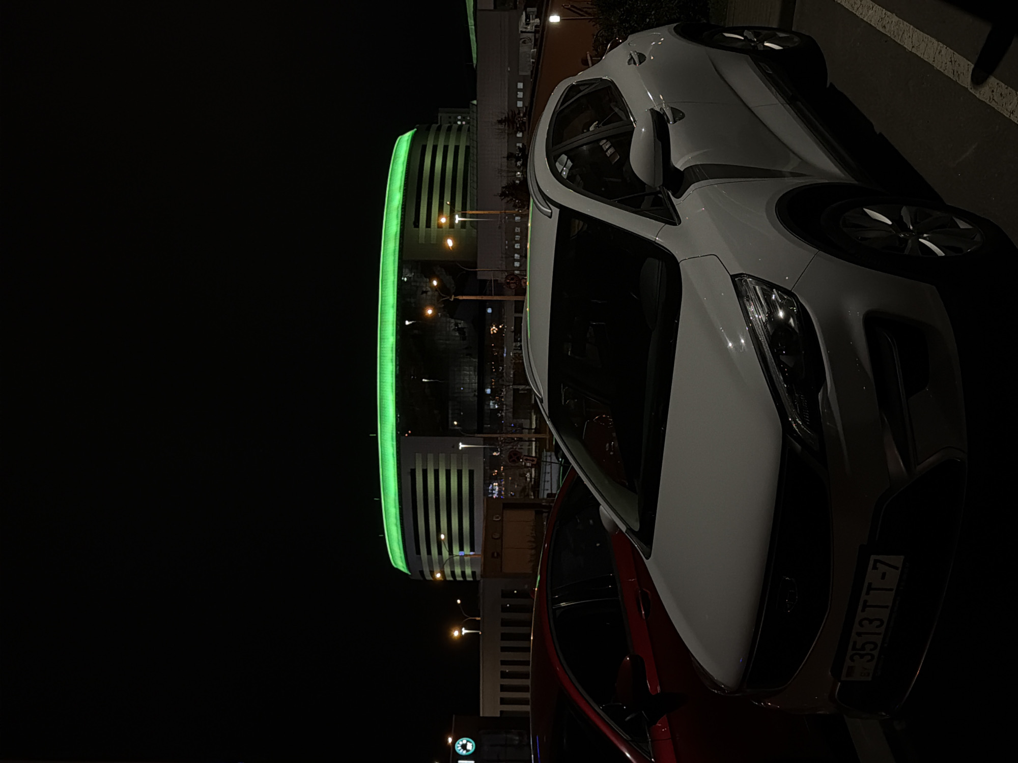 The image displays an urban night scene, dominated by artificial lighting against a dark sky. In the foreground, two cars are parked. A white car, possibly a hatchback or small SUV, is prominently featured on the right, seen from its front-right quarter. Its headlamp and grille are visible, and its front license plate clearly displays the alphanumeric sequence "3513 TT-7" along with the country code "BY", indicating Belarus. A second car, partially visible in a darker red or maroon hue, is parked to the left of the white car.
In the midground, a modern building stands out, characterized by its striking green illumination along its upper structure and horizontal lines on its facade. Around this central building, other less distinct buildings and structures are visible, punctuated by numerous streetlights emitting a warm, yellow-orange glow. The scene is devoid of people, conveying a quiet, nocturnal atmosphere in a city environment, likely a parking lot or street adjacent to commercial buildings.